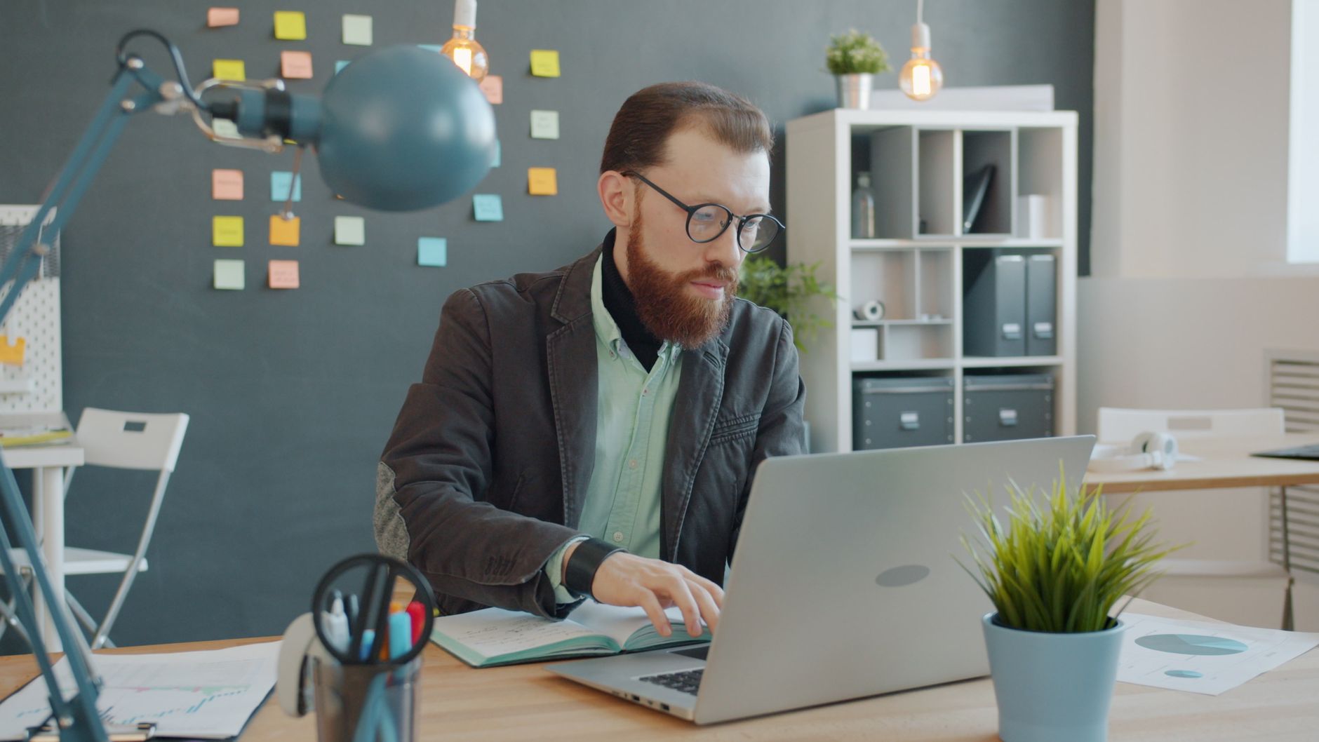 A bearded man wearing glasses types on a laptop at an office desk