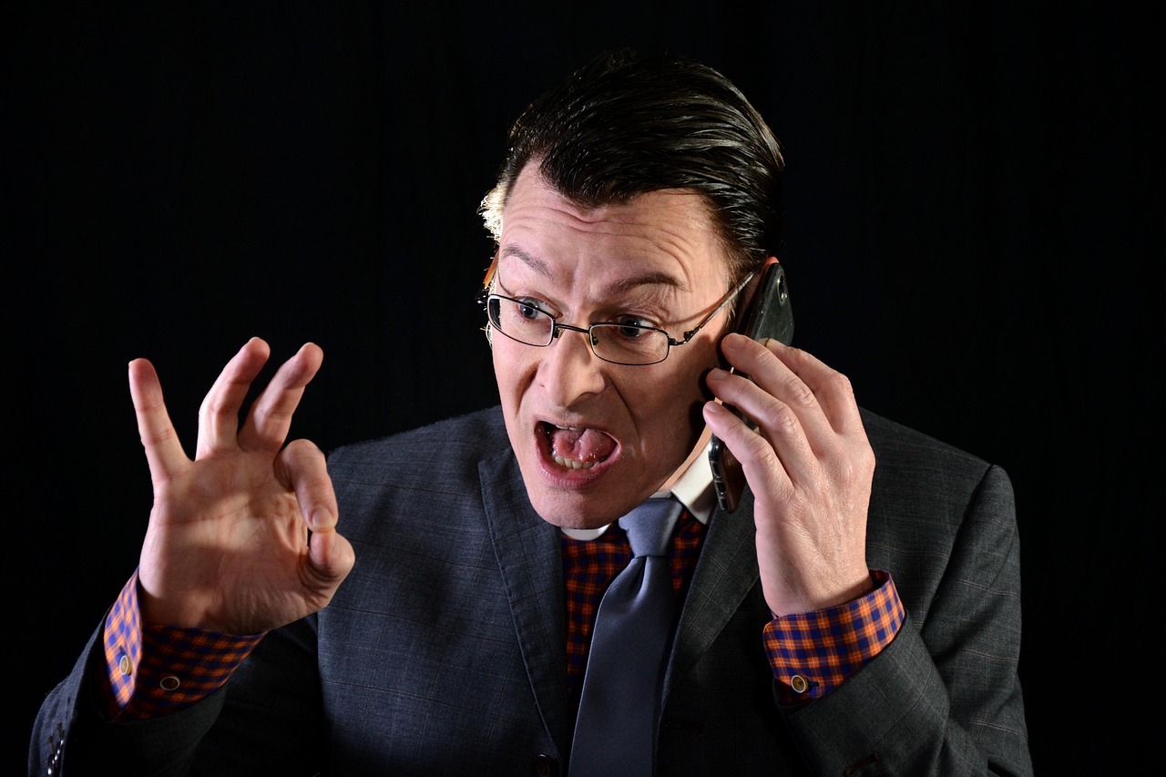 An angry man in glasses talks on the phone, making an "OK" gesture