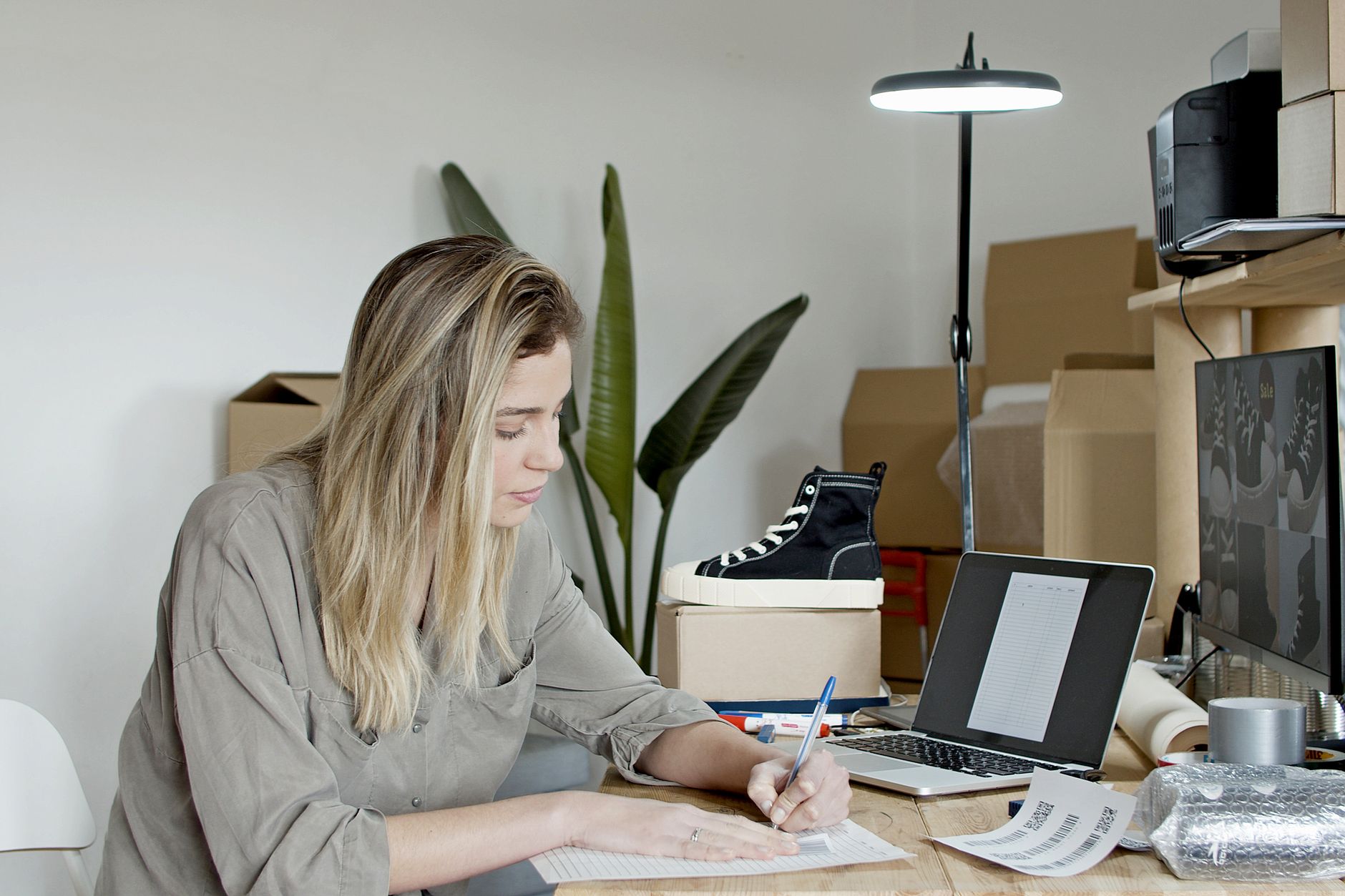 Woman manually writing at a cluttered desk with a laptop and product boxes nearby