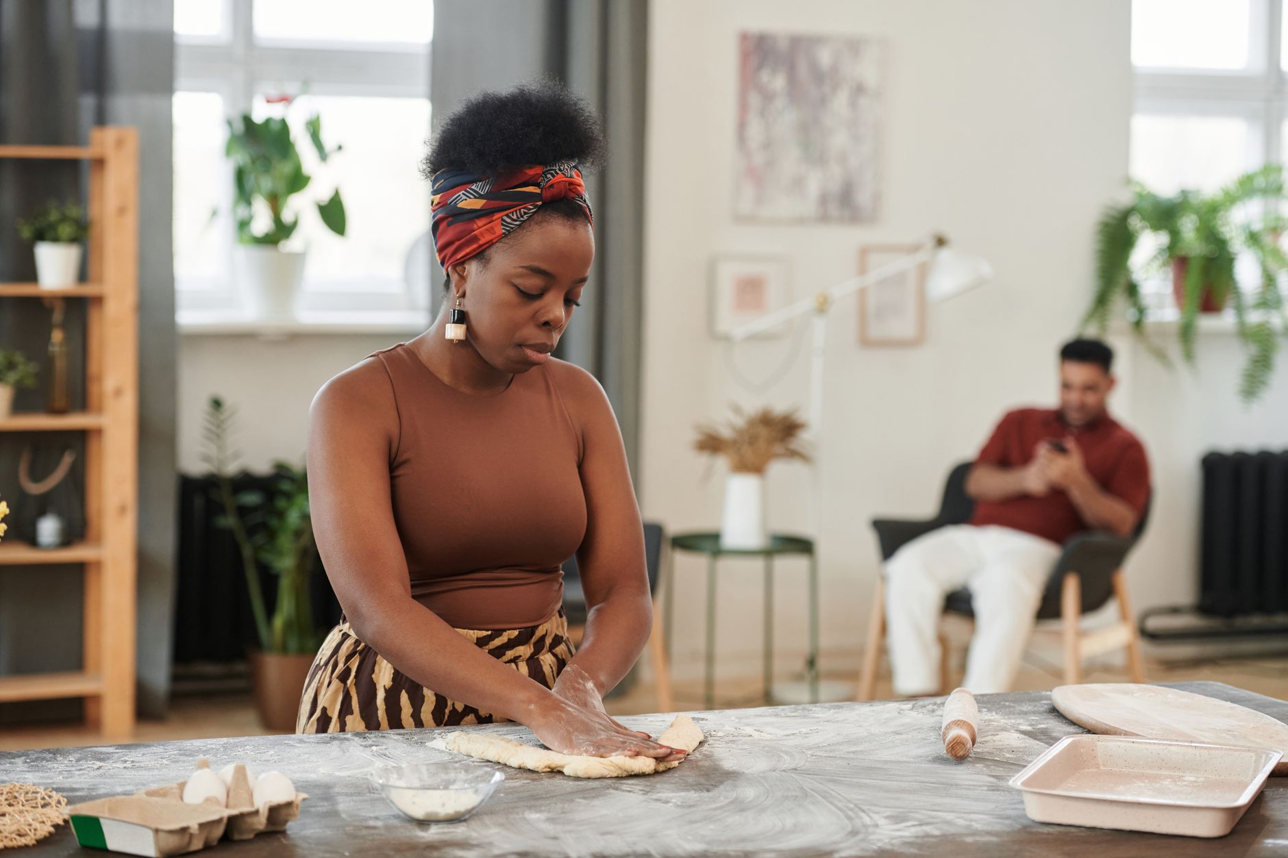 Woman kneading dough on a kitchen counter while a man relaxes in the background