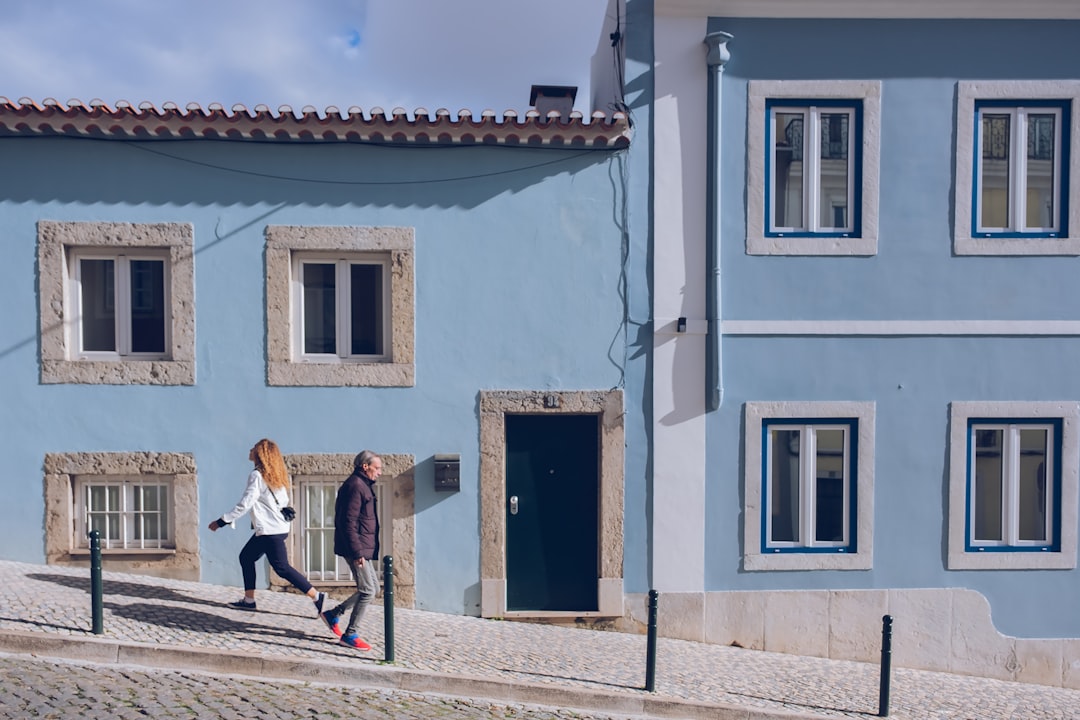 Two people walk uphill on a cobblestone street past light blue buildings in Lisbon