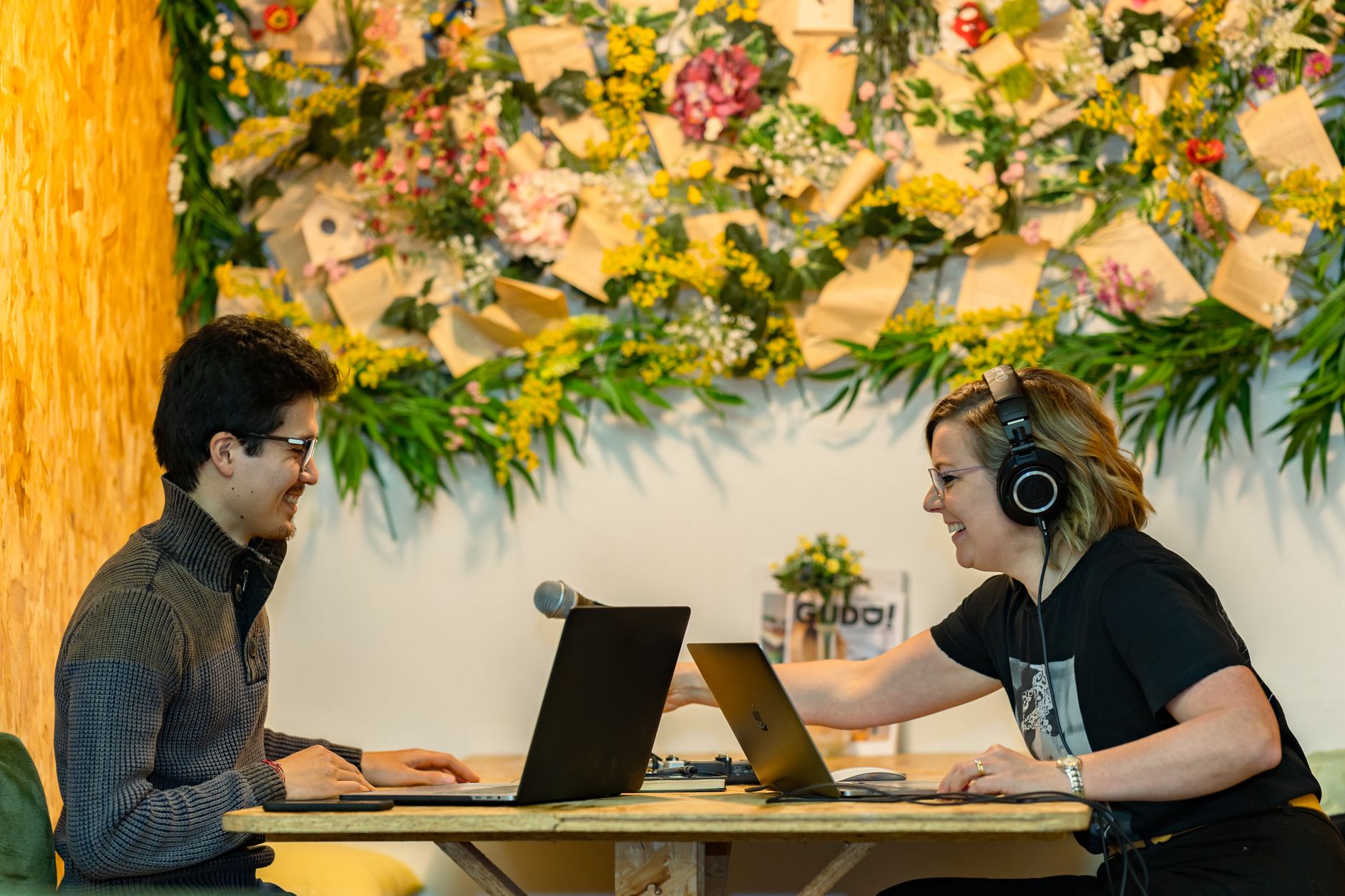 Two smiling people with laptops, headphones, and a microphone, perhaps recording a Bigpond podcast
