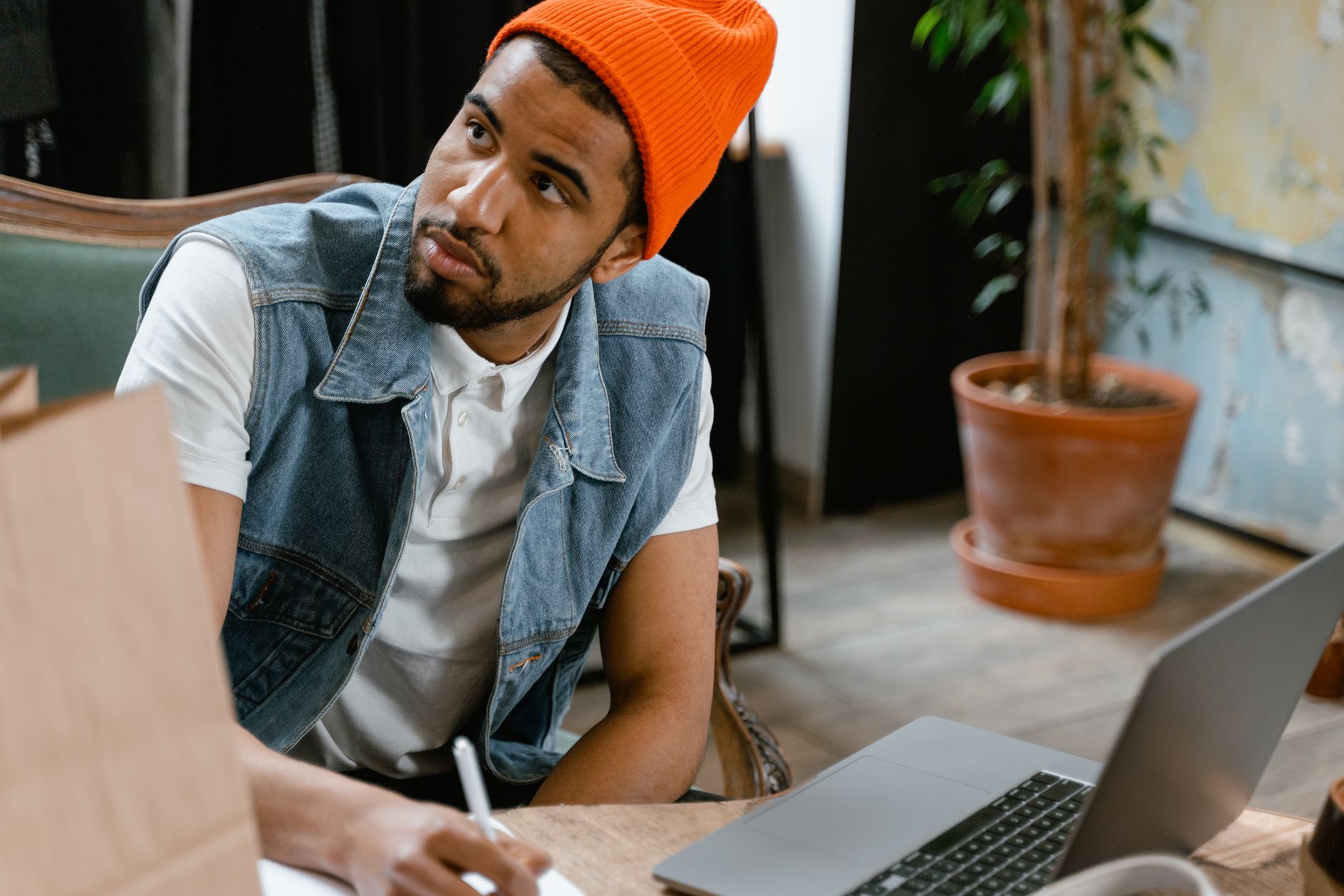 Man in orange beanie and denim vest thinking while writing with laptop nearby