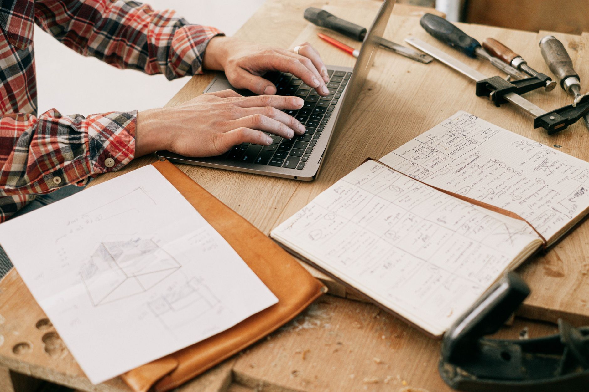 Hands typing on a laptop with a notebook and tools on a wooden workbench