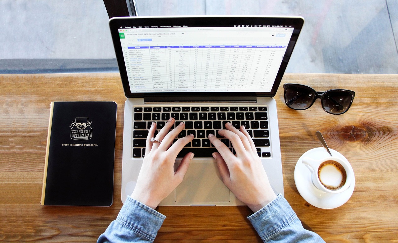 A person types on a laptop displaying a spreadsheet at a wooden desk