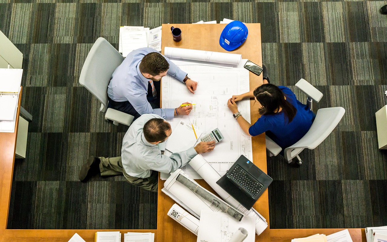 Three people review blueprints and calculations at a large conference table