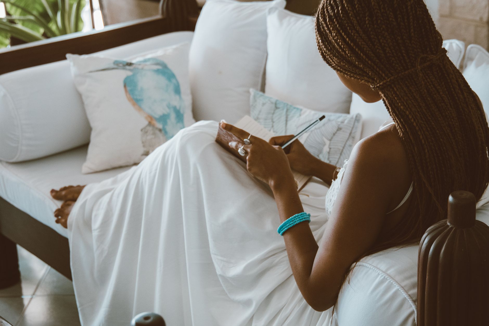Woman with braided hair writing in a journal on a white sofa indoors