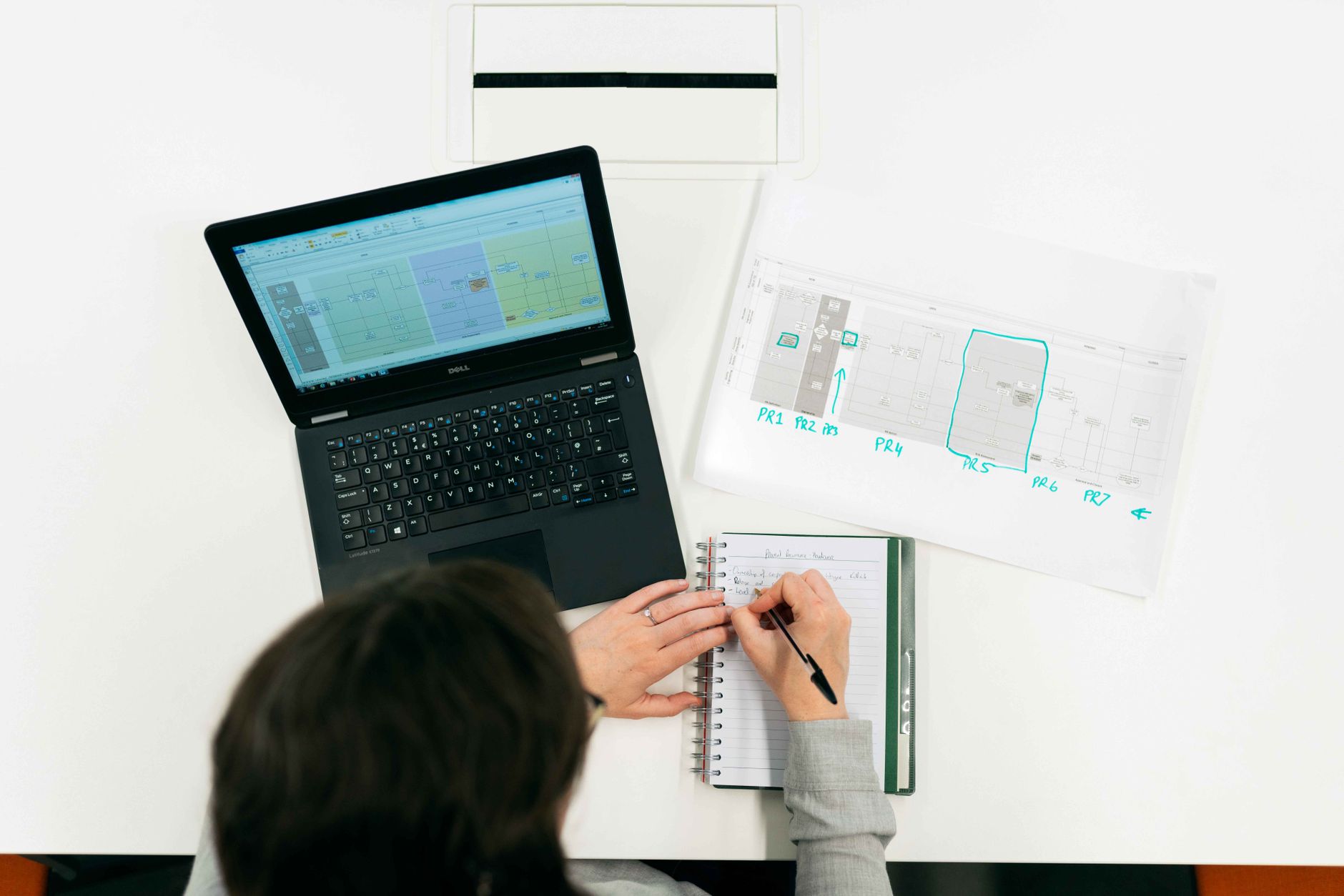 A person writes in a notebook beside a laptop on a white desk