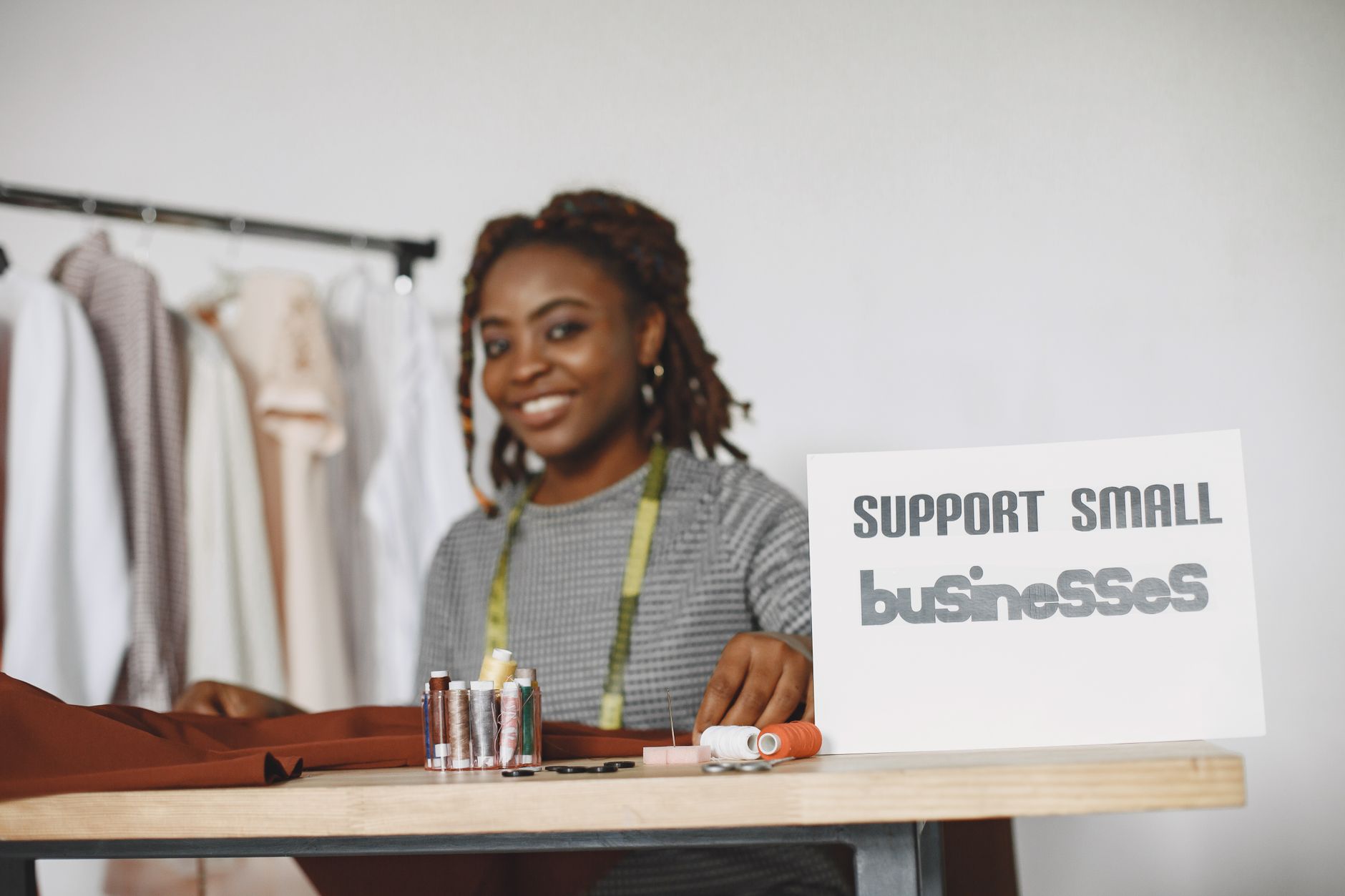 Smiling Black woman seamstress with a "Support Small Businesses" sign and sewing supplies