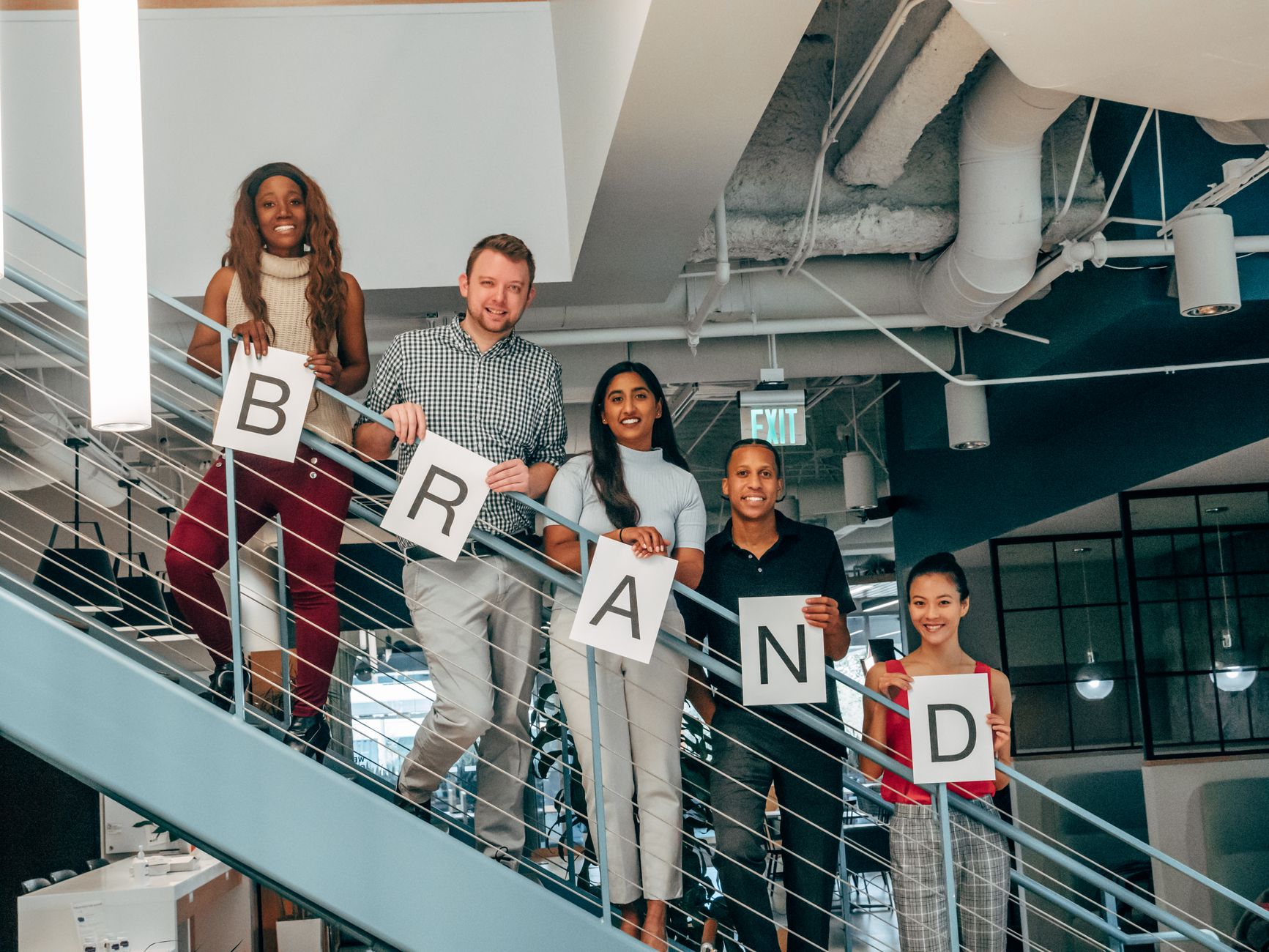 Five diverse people on a modern staircase holding signs spelling out "BRAND" for digital solutions
