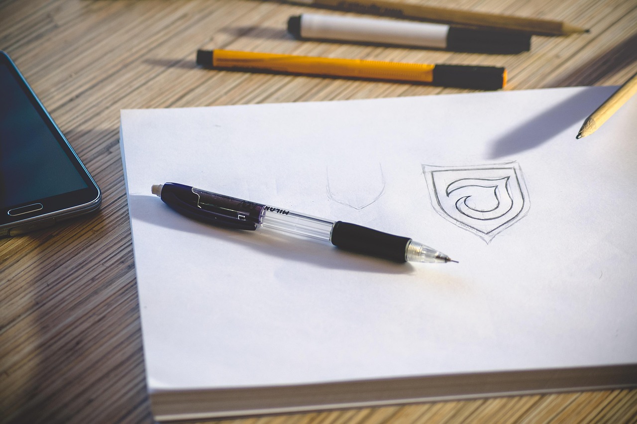 A drawn logo sketch on paper with a pen, phone, and pencils nearby
