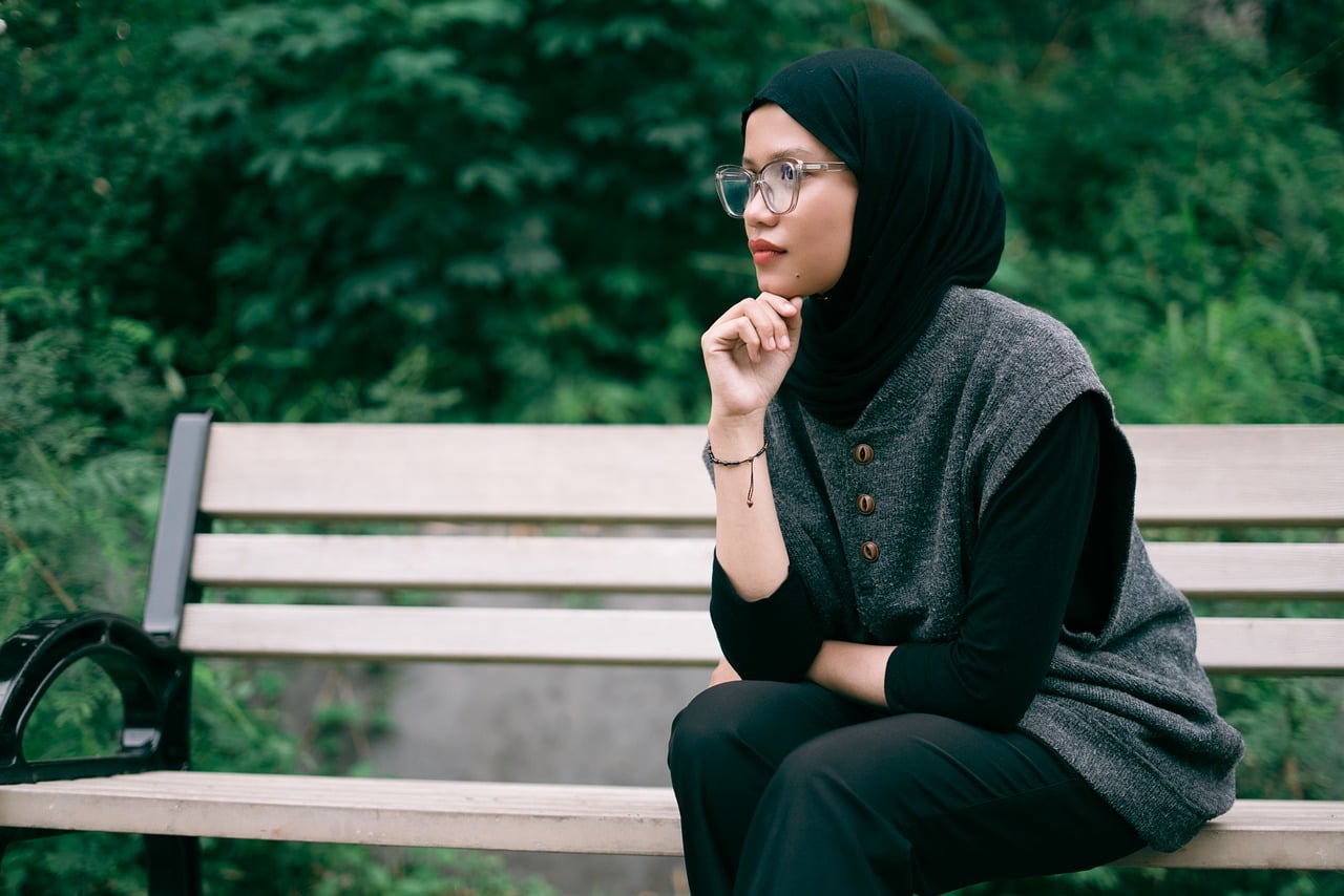 Woman in hijab and glasses sits reflectively on a bench amidst green foliage