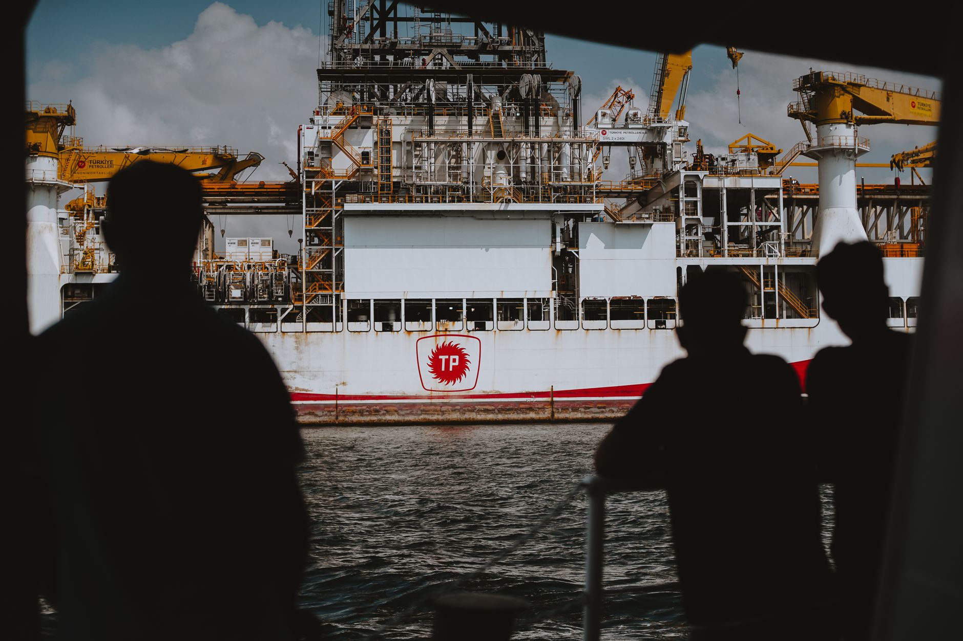Silhouetted people view a large drilling ship with a "TP" logo on the water