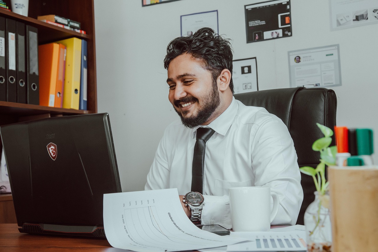 Smiling man in white shirt working on laptop with financial documents at a desk