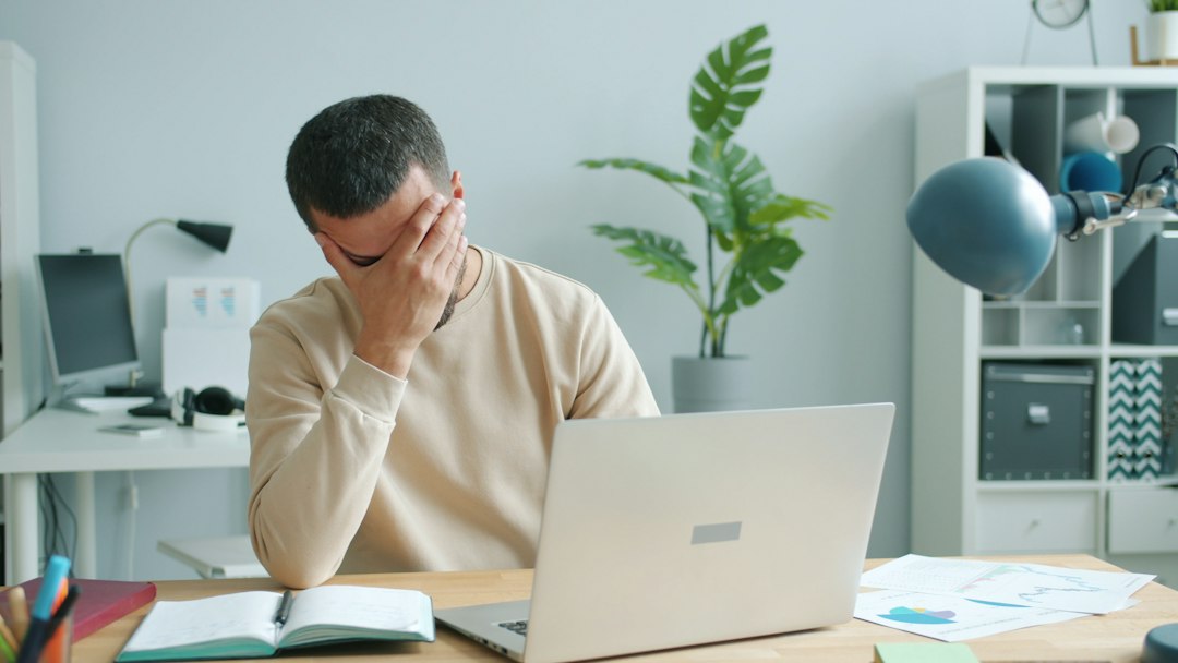 A man sitting before an open laptop covers his face with his hand