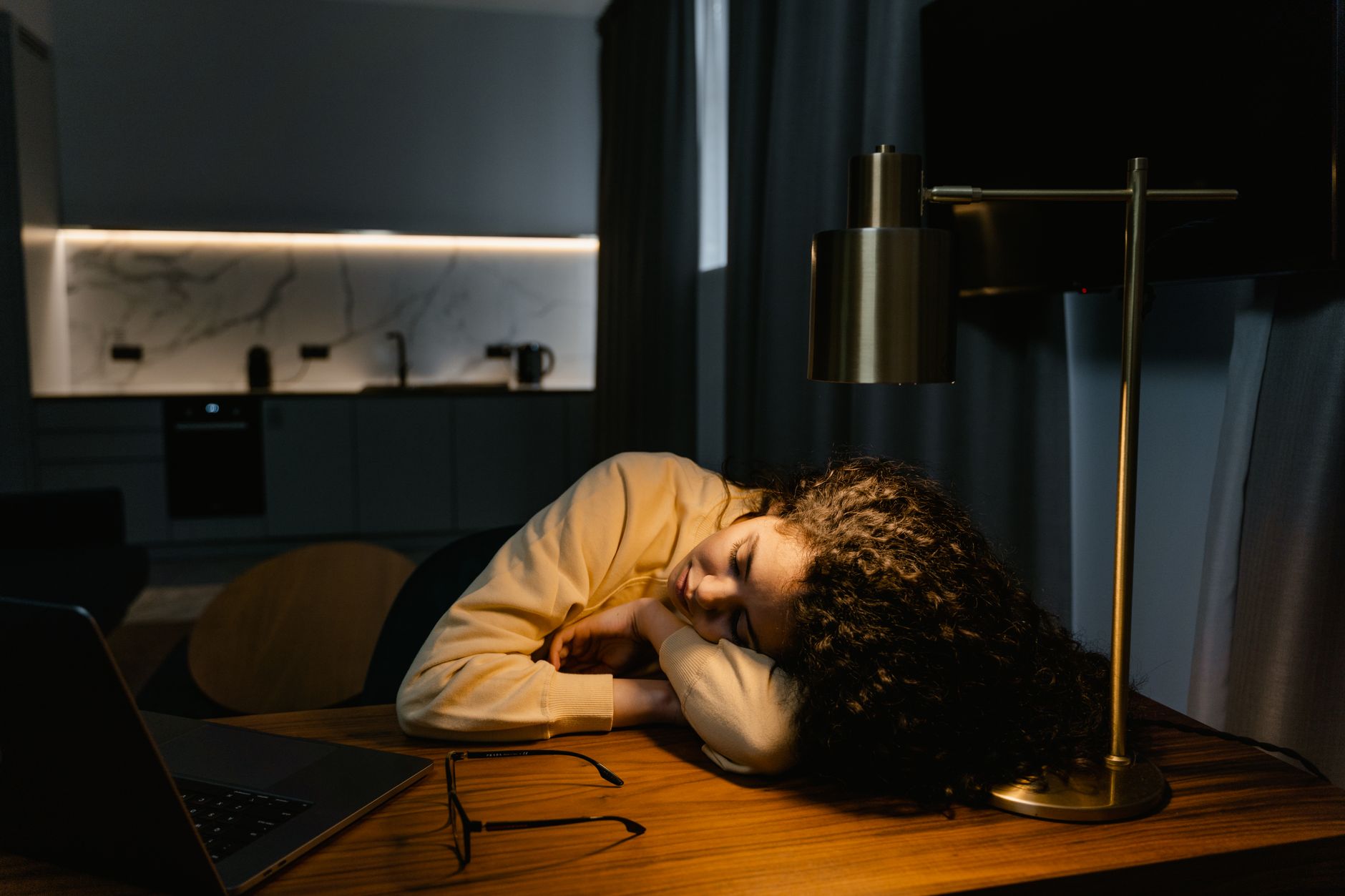 A tired woman with curly hair sleeps on a desk beside a laptop and lamp