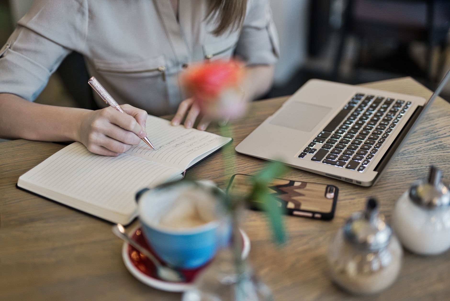 Person writes in notebook next to laptop and coffee, illustrating human-AI collaboration