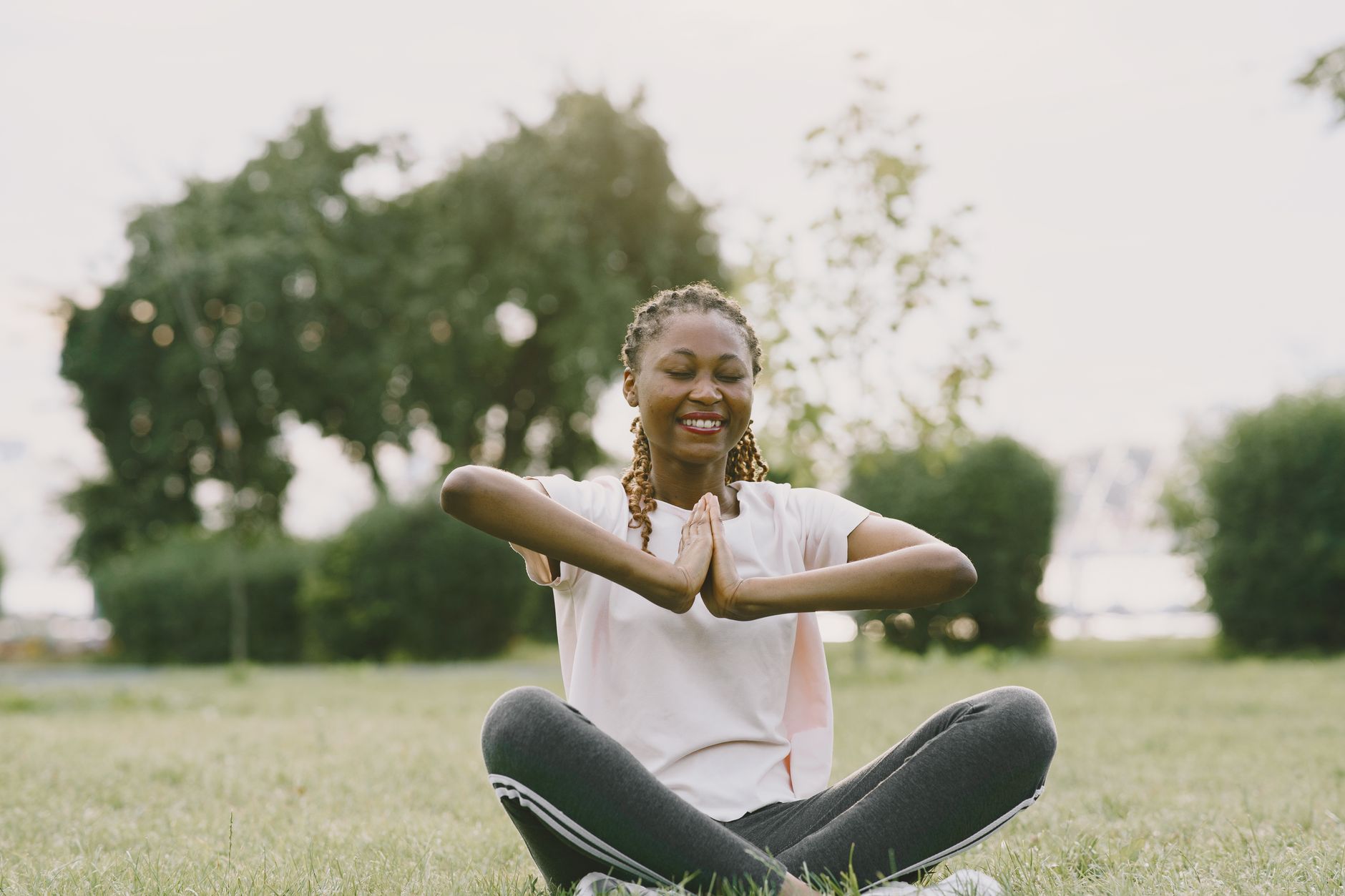 Smiling woman with braided hair meditating in a grassy park setting