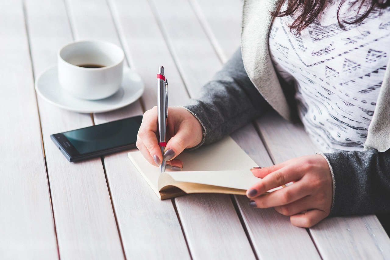 Person writing in a notebook, with coffee and phone on a white table
