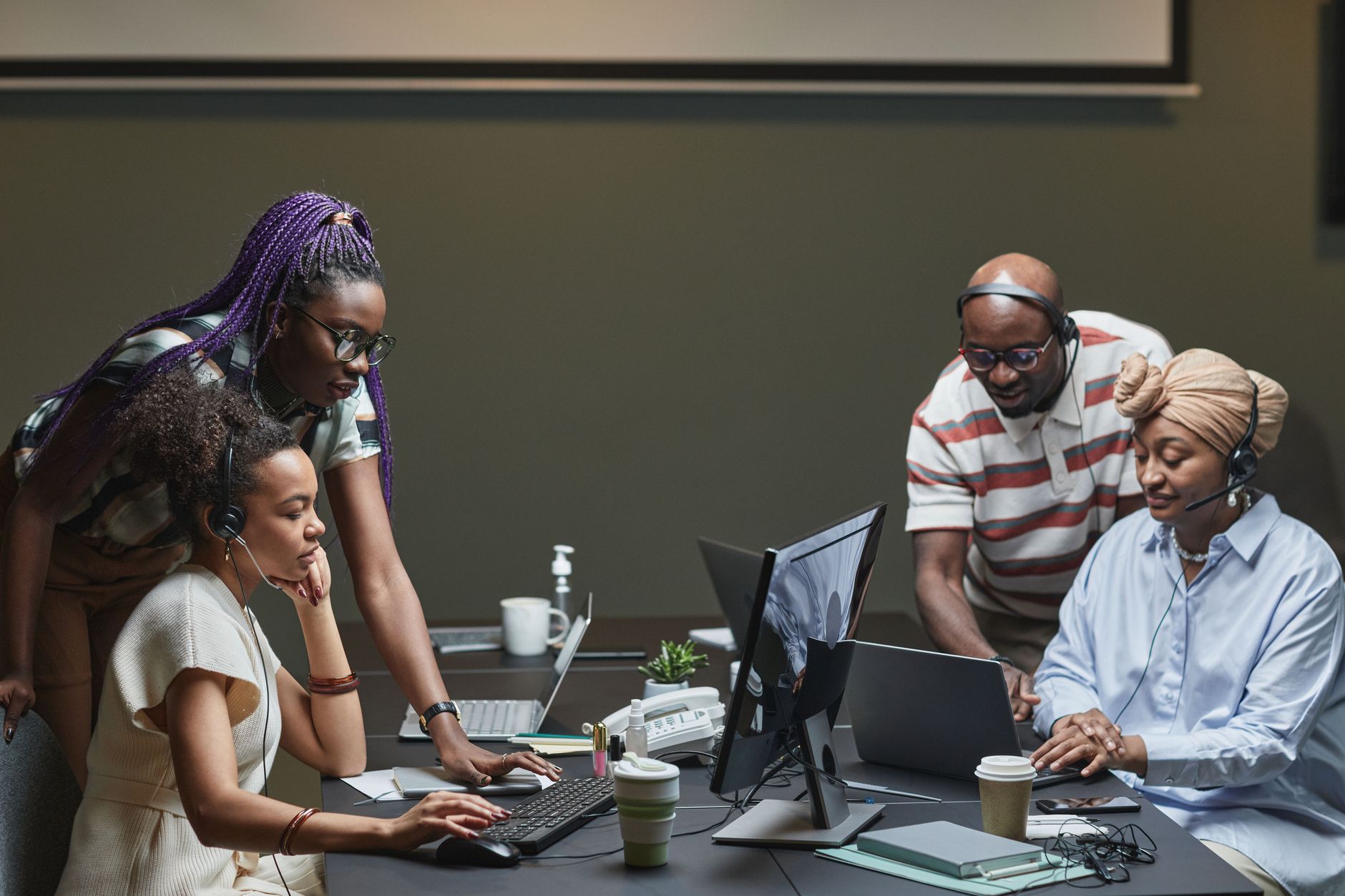 Four diverse people collaborate on computers, wearing headsets in an office environment