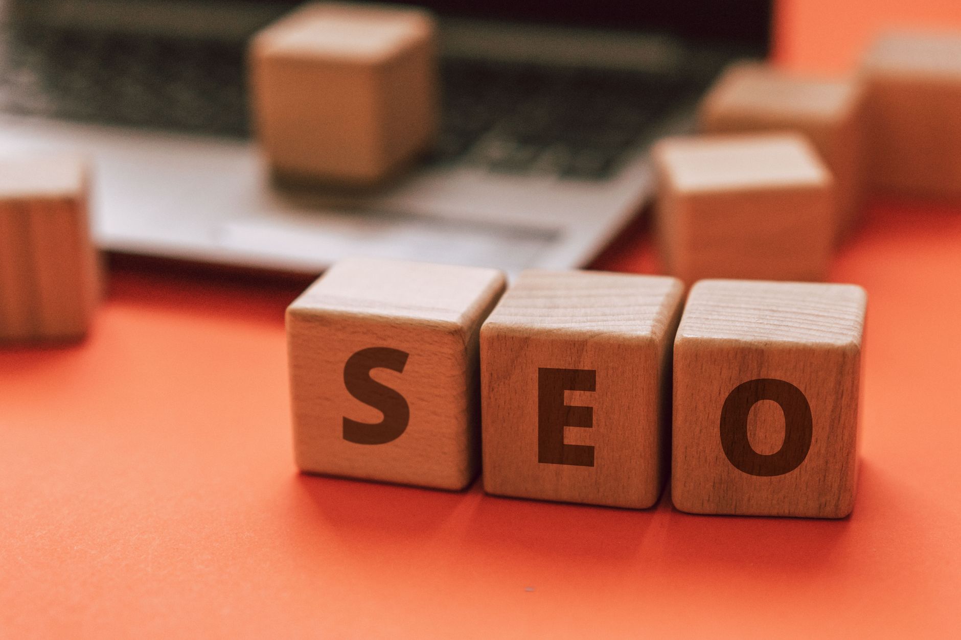 Wooden blocks spell SEO on an orange desk with a laptop visible behind them