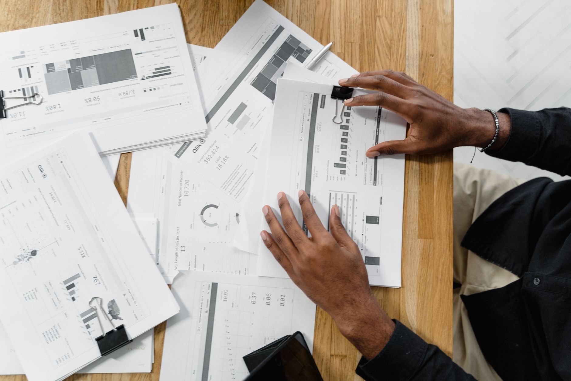 Hands rest on printed charts and documents scattered across a wooden table