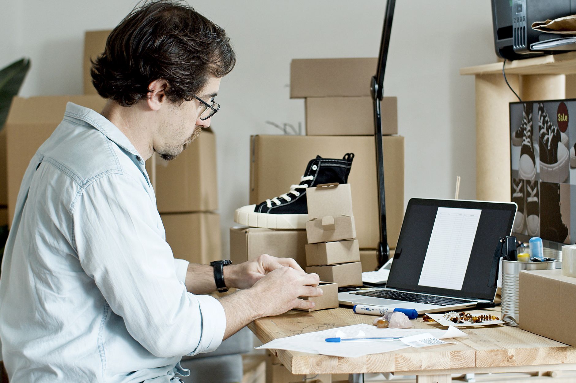 A man sits at a desk packing small cardboard boxes beside a laptop
