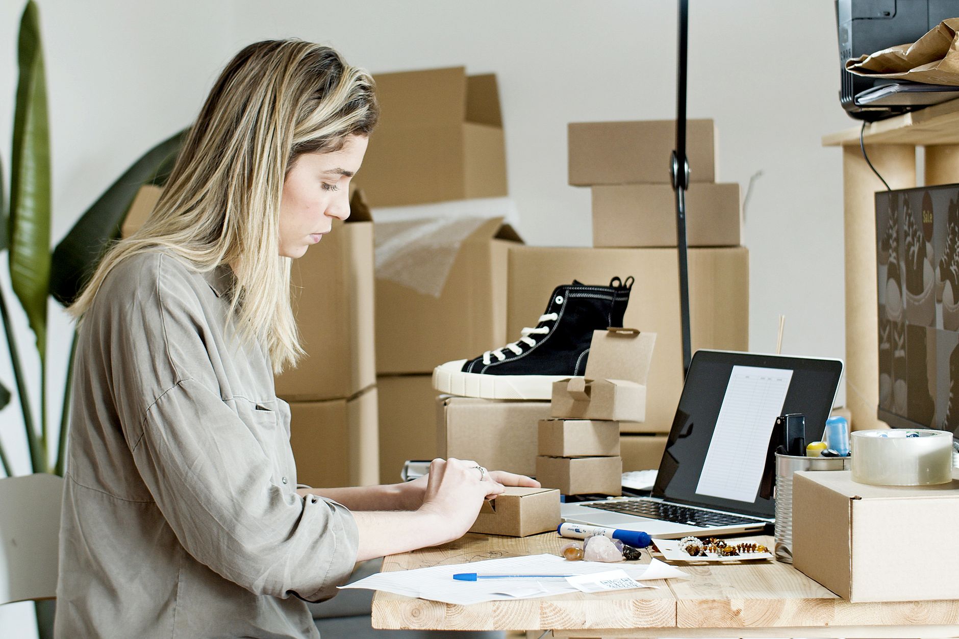 A woman packs a small cardboard box at a desk beside a laptop