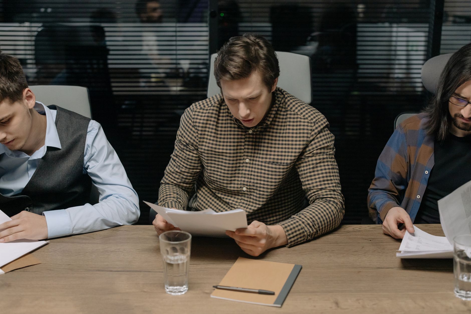 Three men sit at a wooden conference table reading printed paper documents