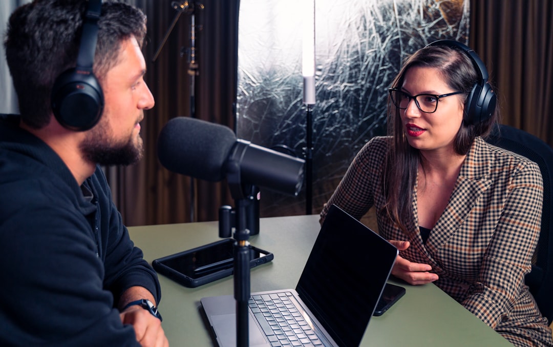 Two people wearing headphones sit at a table with a microphone and laptop, podcasting