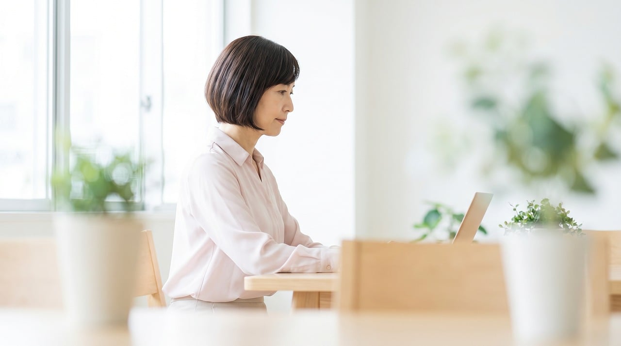 A woman sits at a wooden table using a laptop near indoor plants