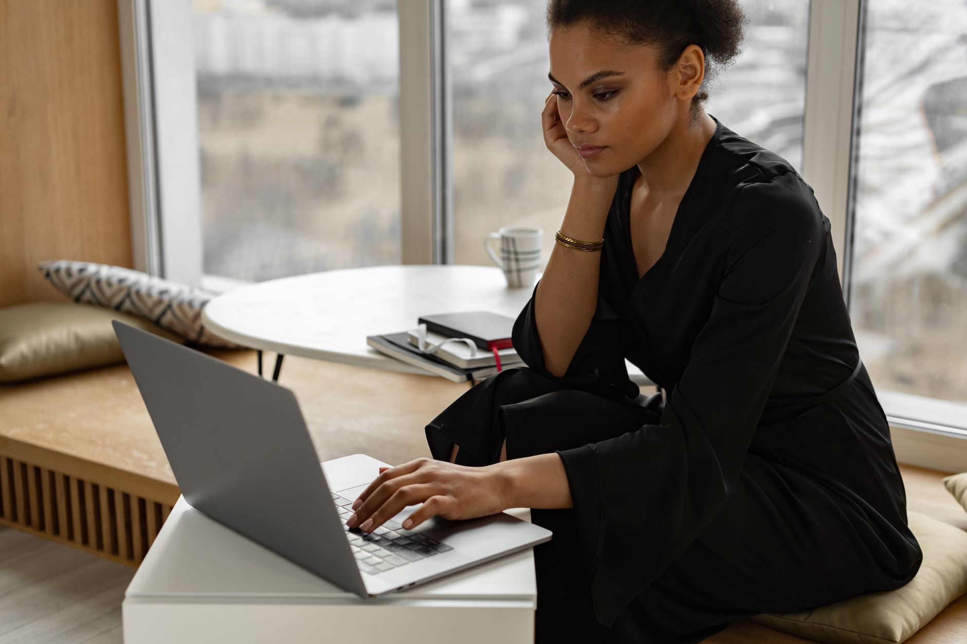 A woman in a black dress types on a laptop beside a window