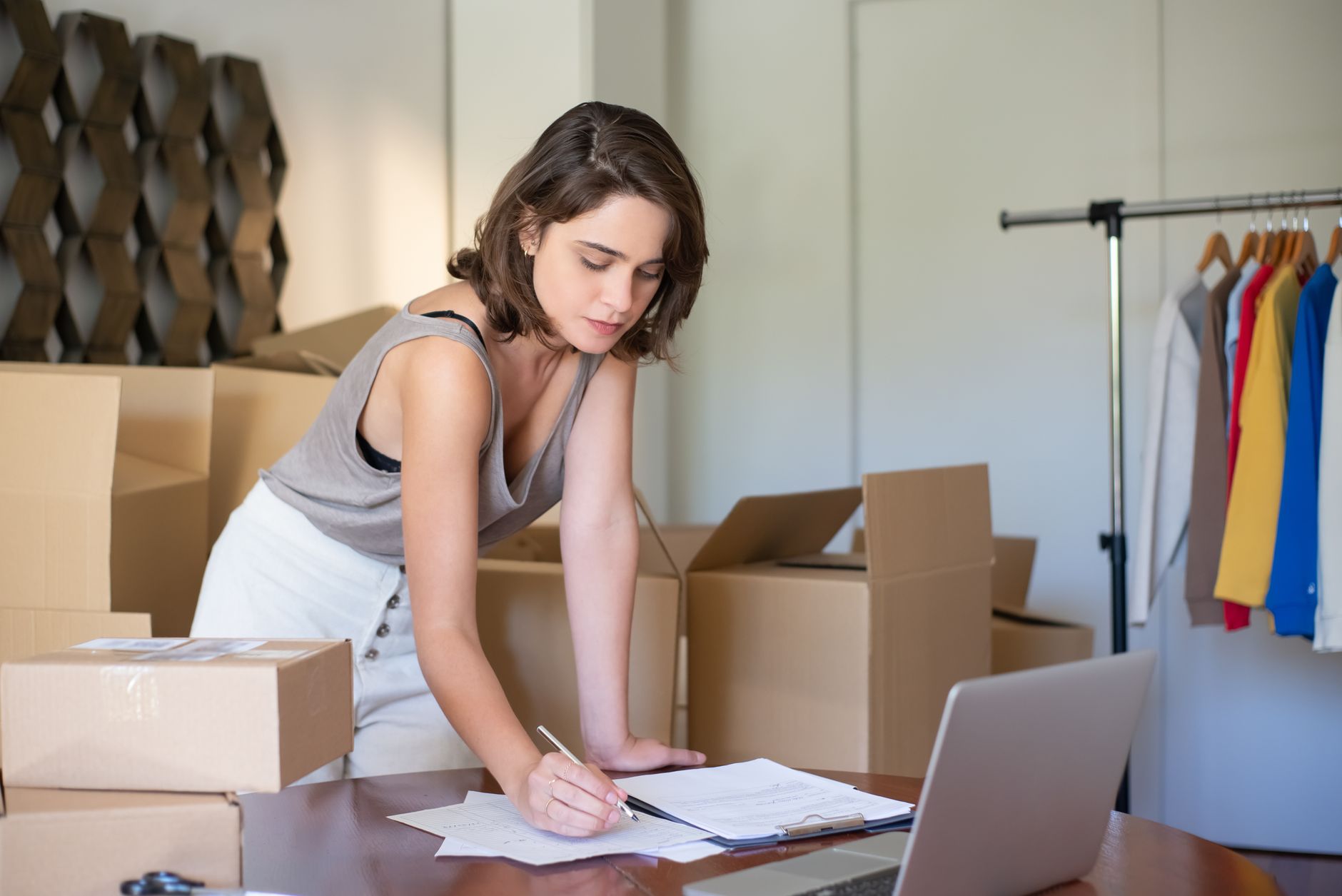 Woman working on paperwork at a desk with boxes and clothes rack nearby