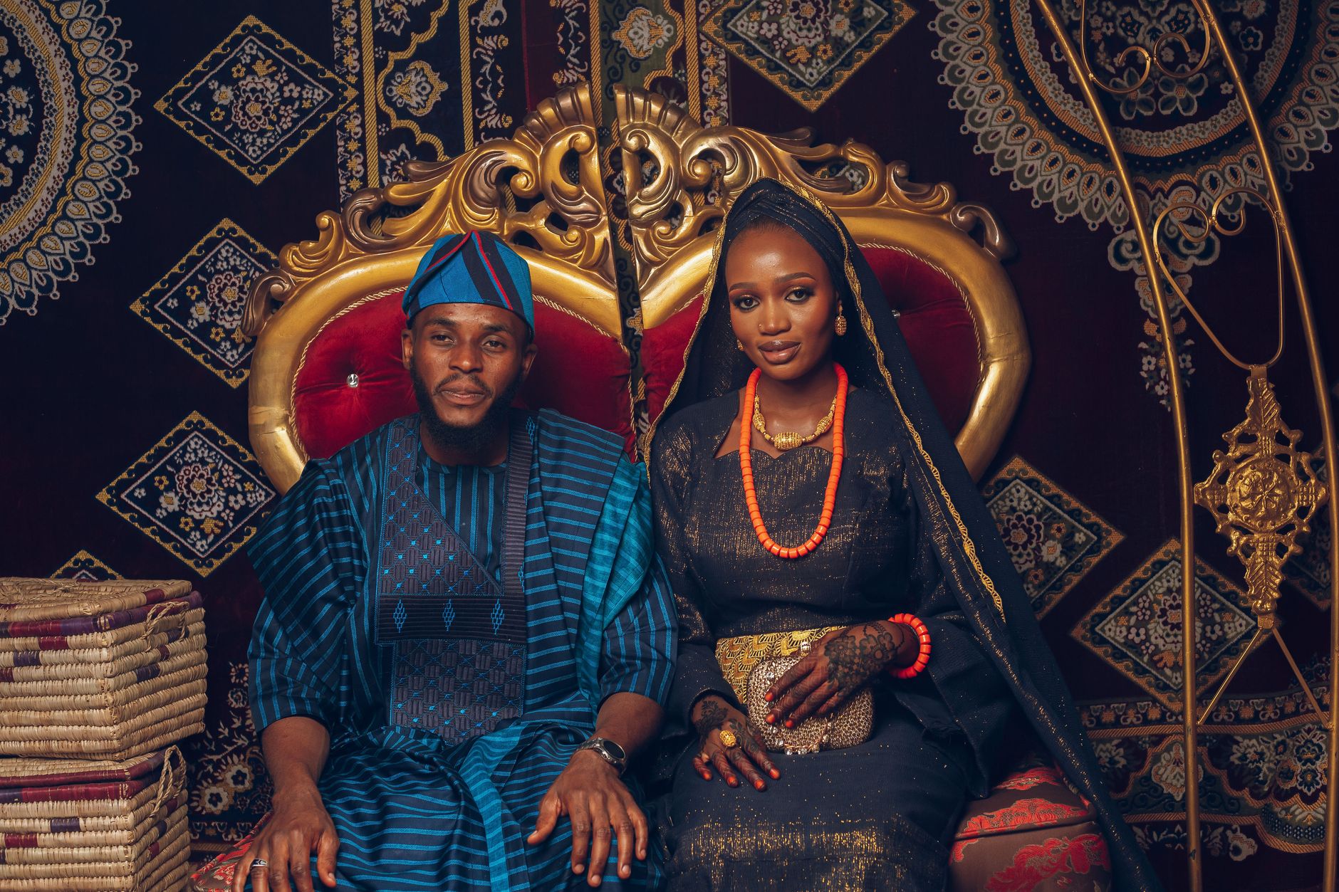 A Nigerian couple in traditional attire sits on an ornate gold and red wedding throne