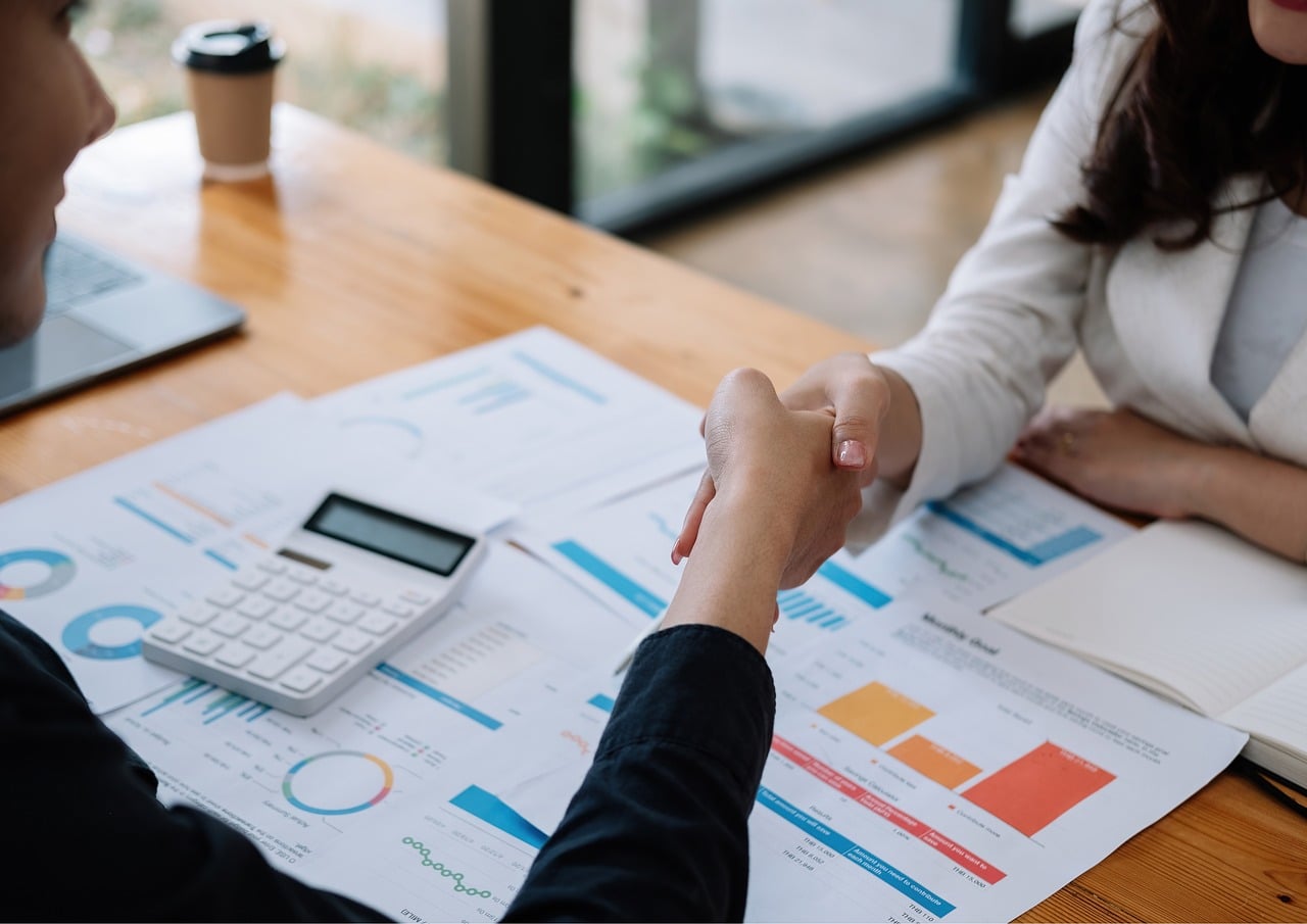 Two people shake hands over financial charts and a calculator during a business consultation