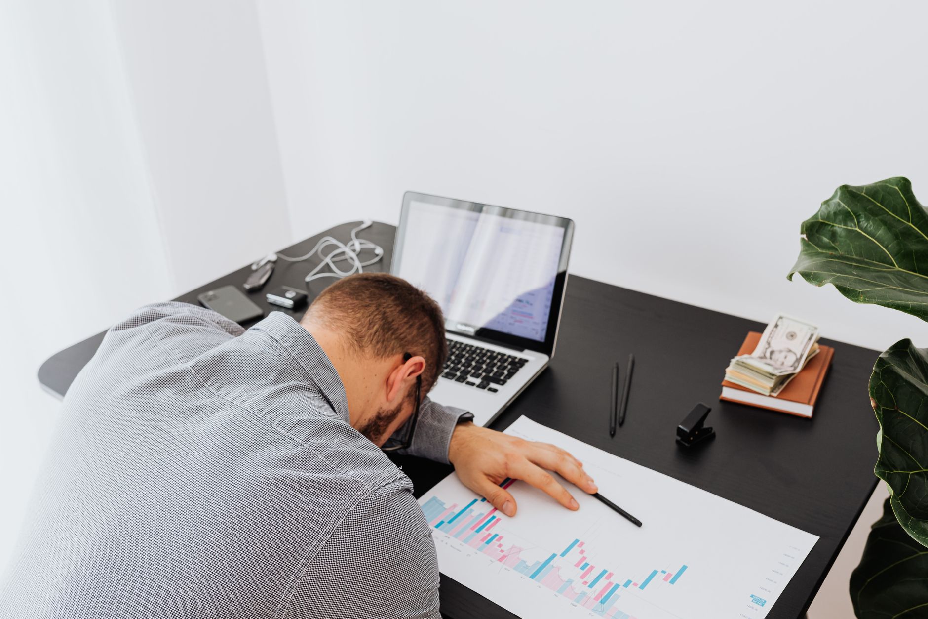 A man rests his head on a desk beside charts and a laptop