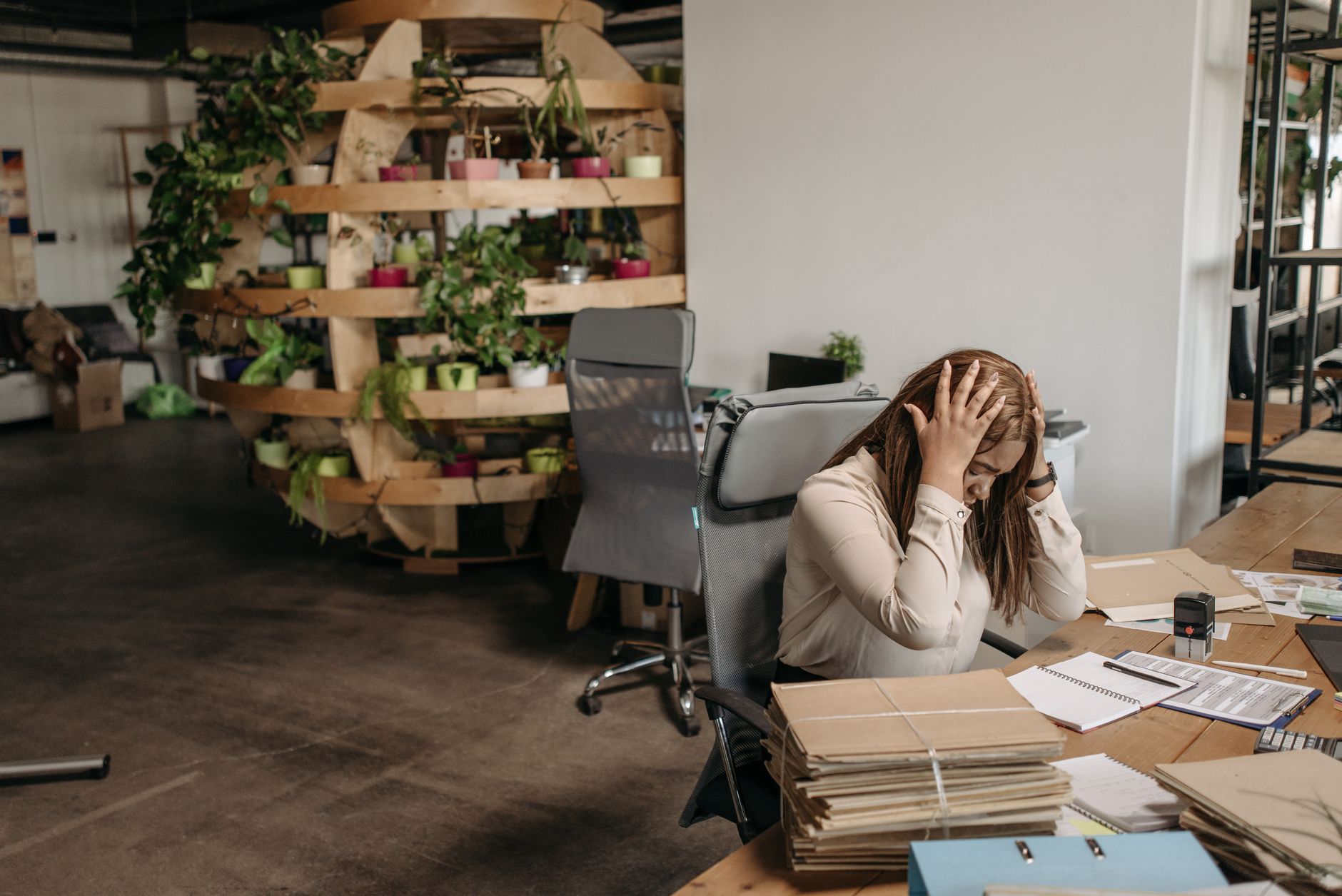 A woman holding her head sits at a desk beside stacks of paper