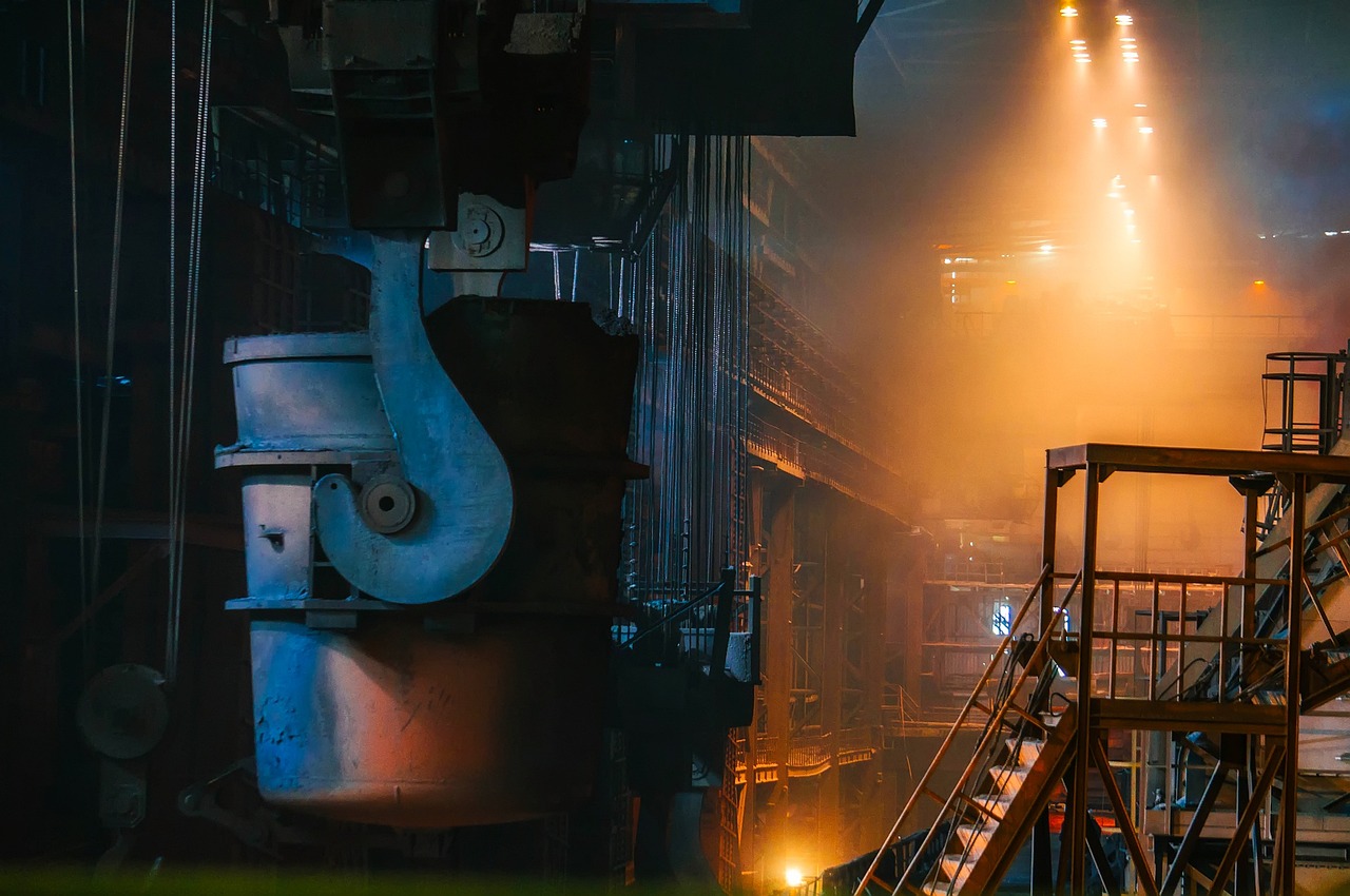 A large industrial crane hook and ladle against a smoky, brightly lit factory interior