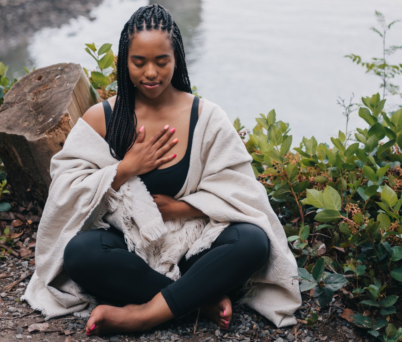 Woman meditating outdoors by water, hands placed on her chest for mindfulness practice