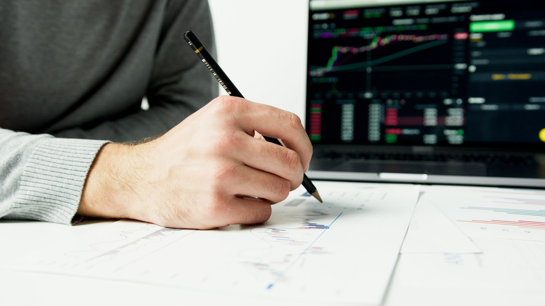 Hand uses pencil to analyze charts on paper, with more analytics visible on laptop