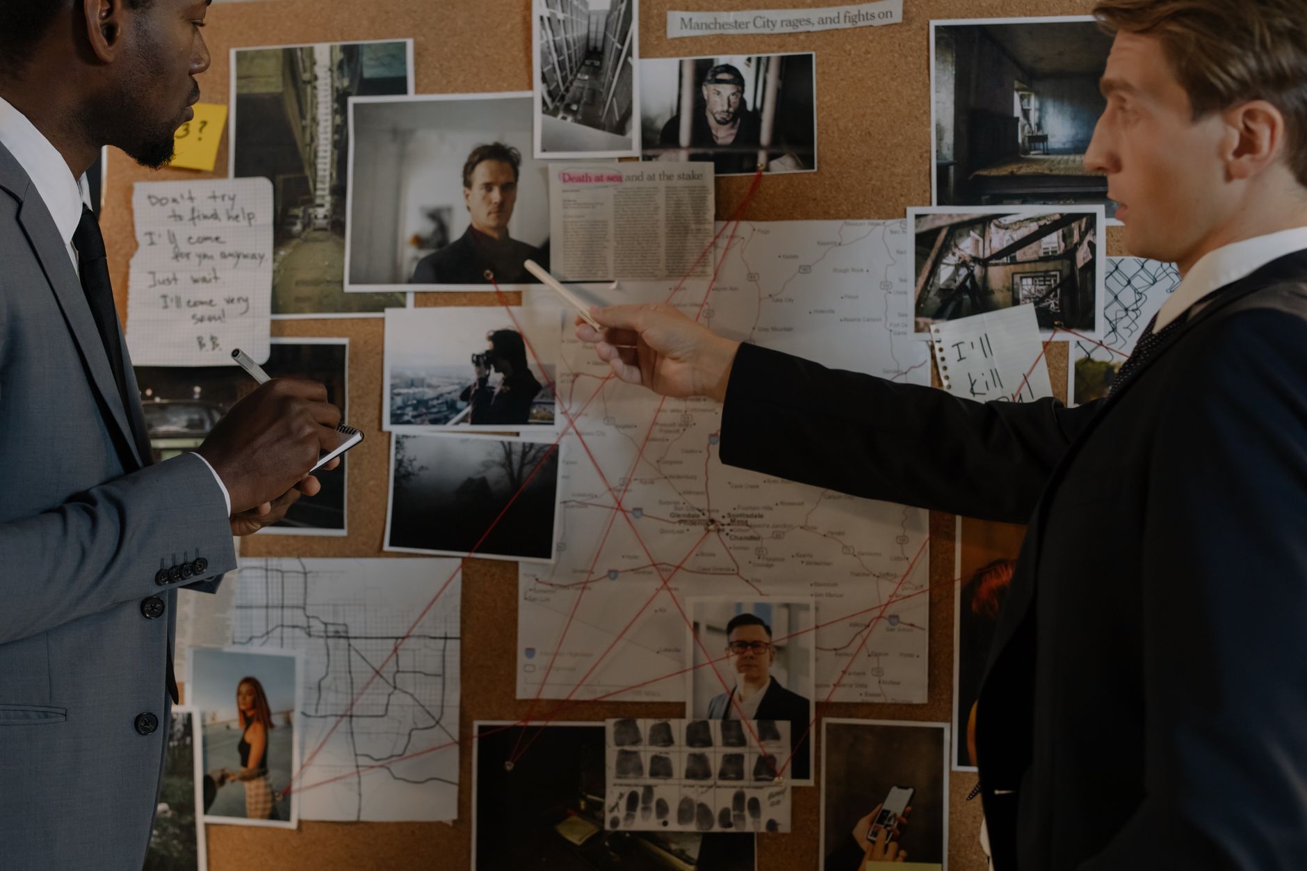 Two men investigate a cork board covered with photos, maps, and notes