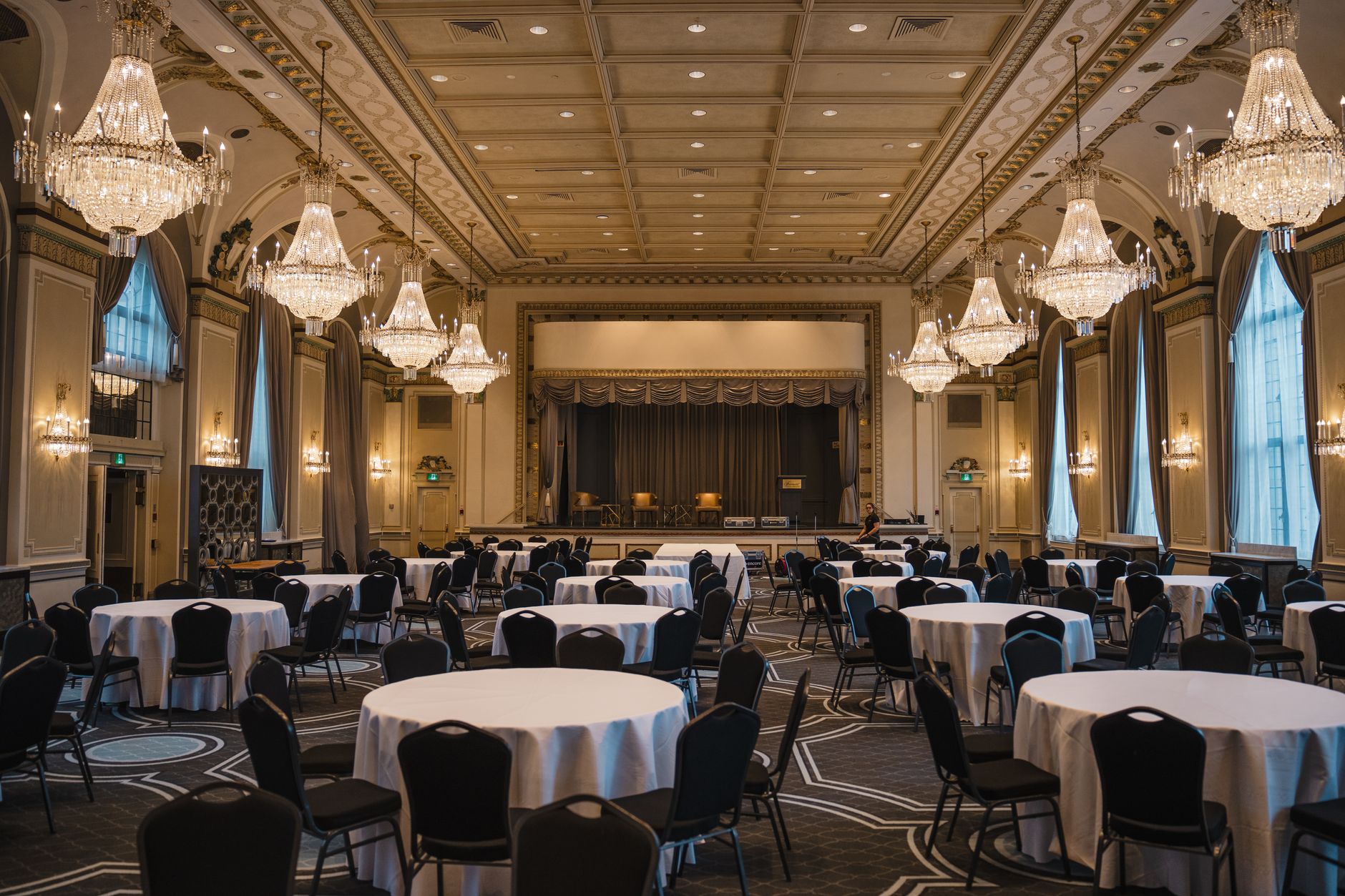 A grand ballroom with round tables and chairs, under many crystal chandeliers