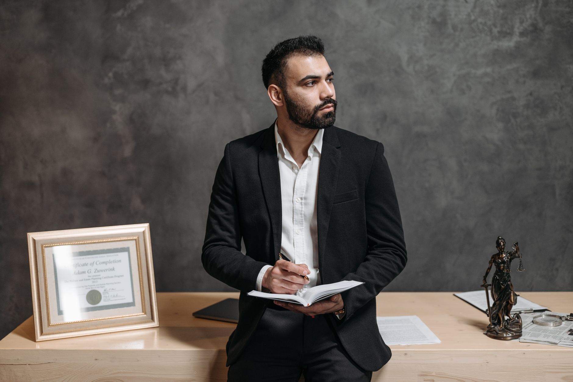 A lawyer in a suit holds a notebook, with certificates and Justice statue nearby