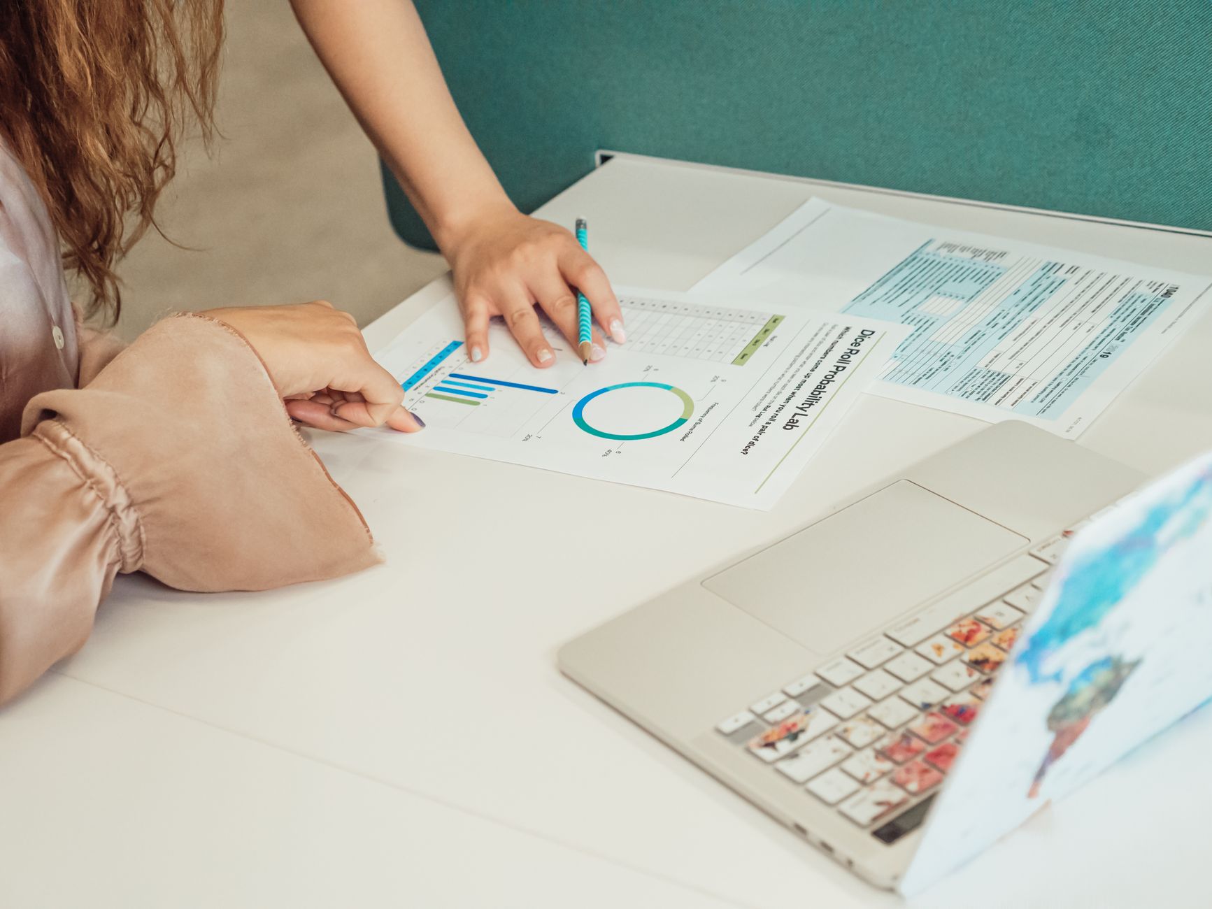 Two people analyze data charts, forms, and a laptop on a white desk
