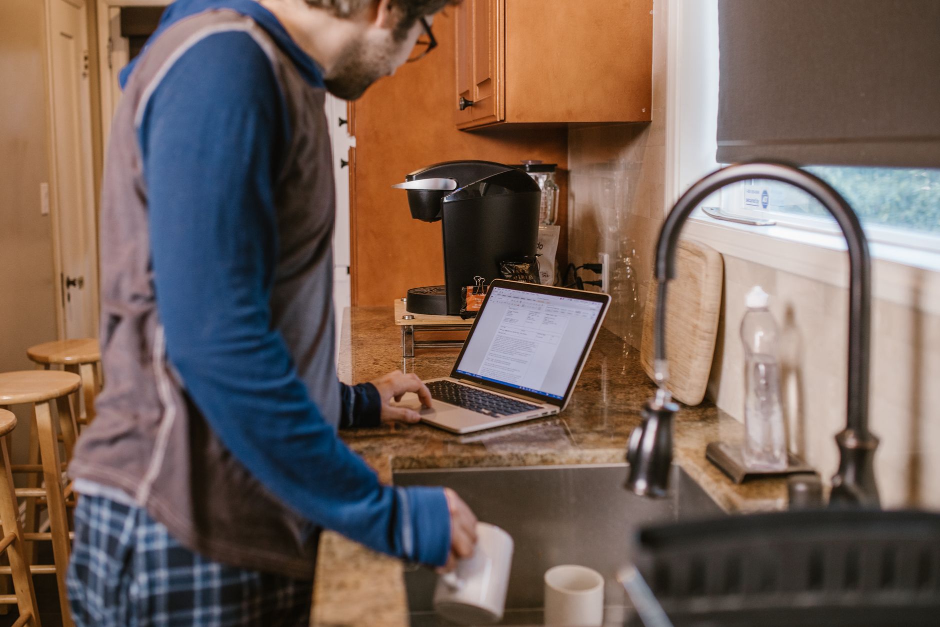 Man in kitchen working on laptop while holding a coffee mug, casual work-from-home setting