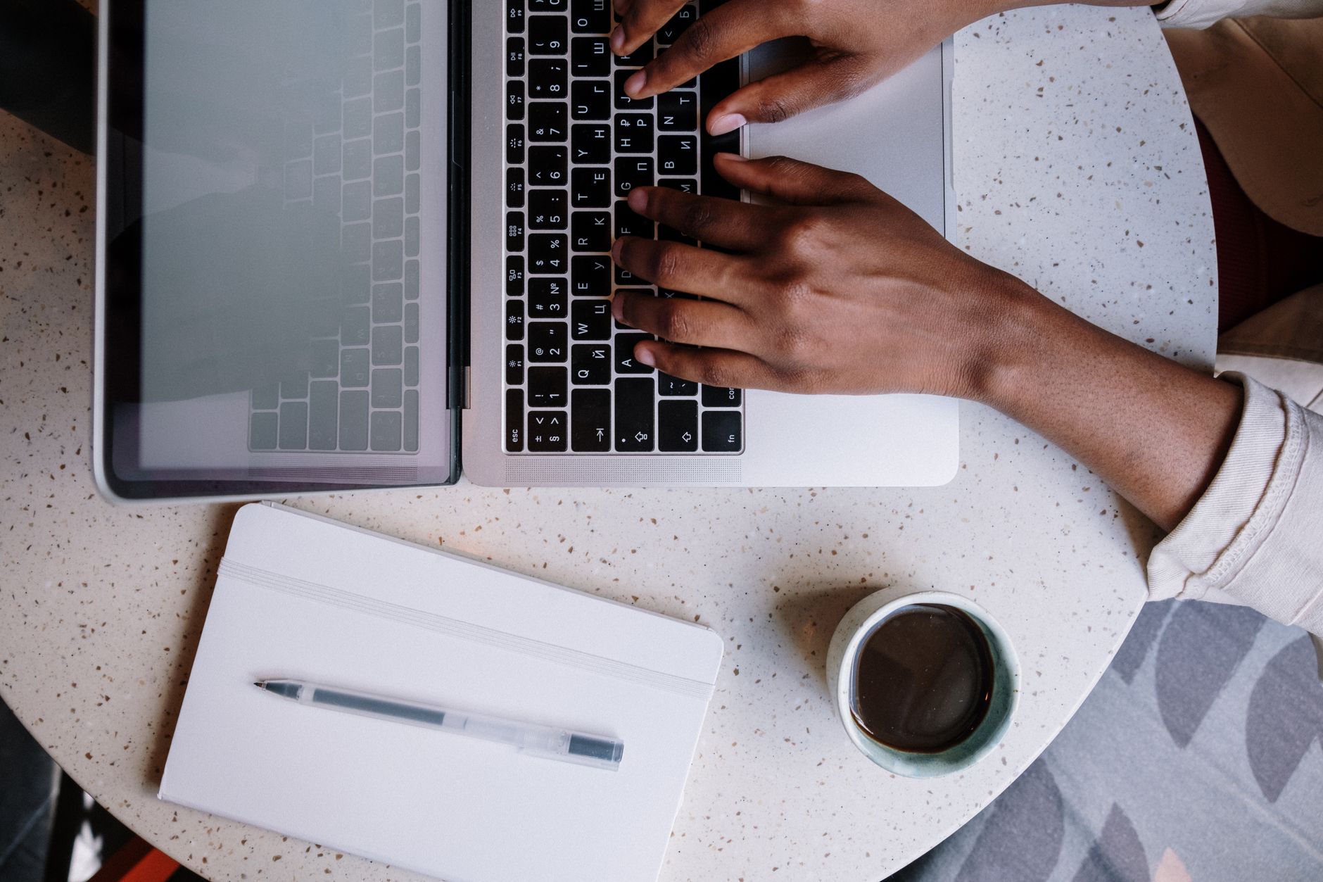 Hands type on a laptop keyboard, next to a notebook and coffee cup