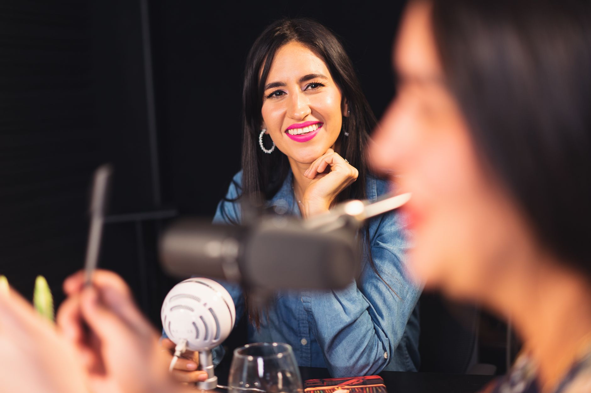 A smiling woman at a podcast recording table with microphones, engaging in conversation