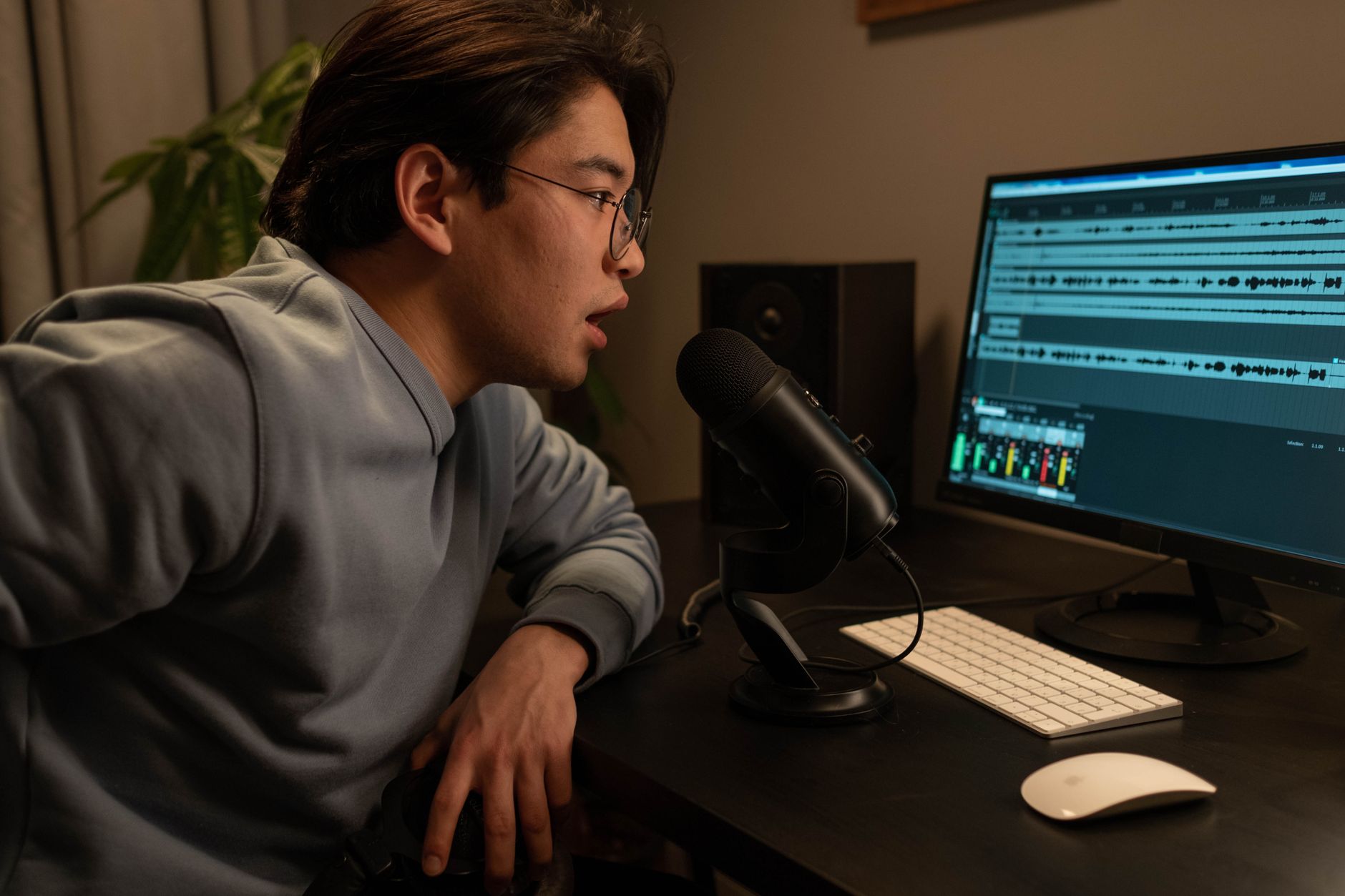 A man speaks into a microphone beside a monitor displaying audio editing software