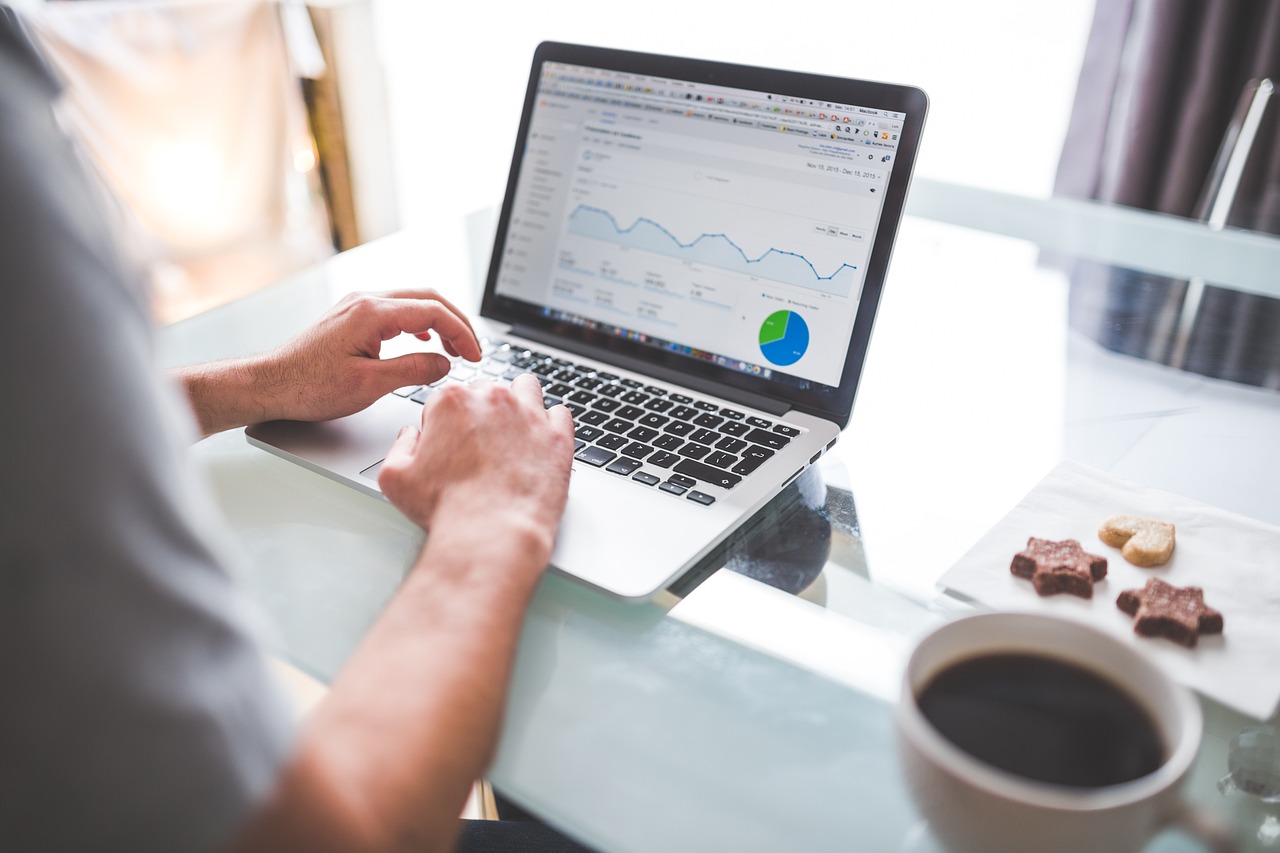 A person types on a laptop displaying analytic graphs next to coffee and cookies