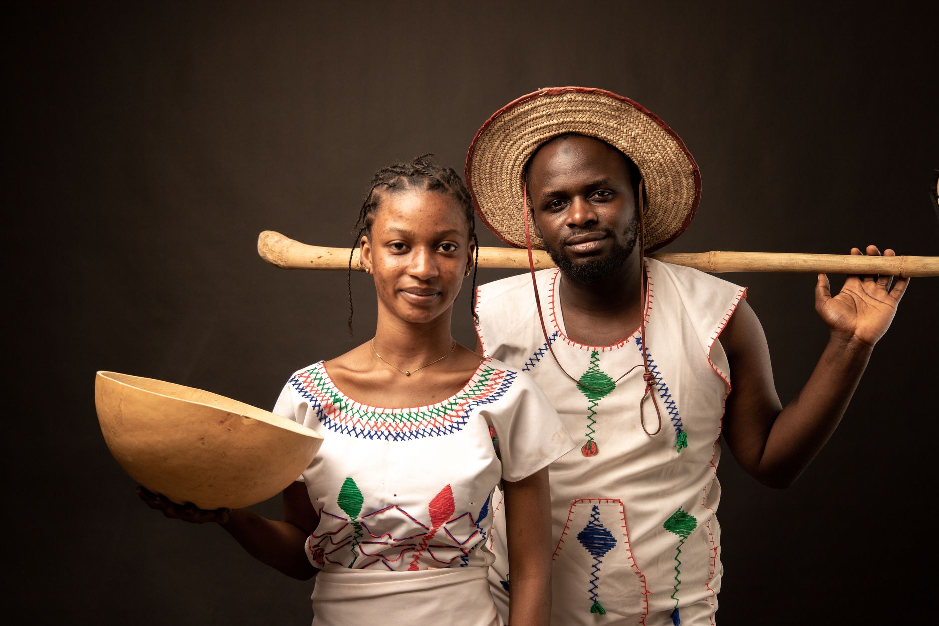 A Nigerian couple in traditional attire, the man wearing a straw hat and holding a hoe